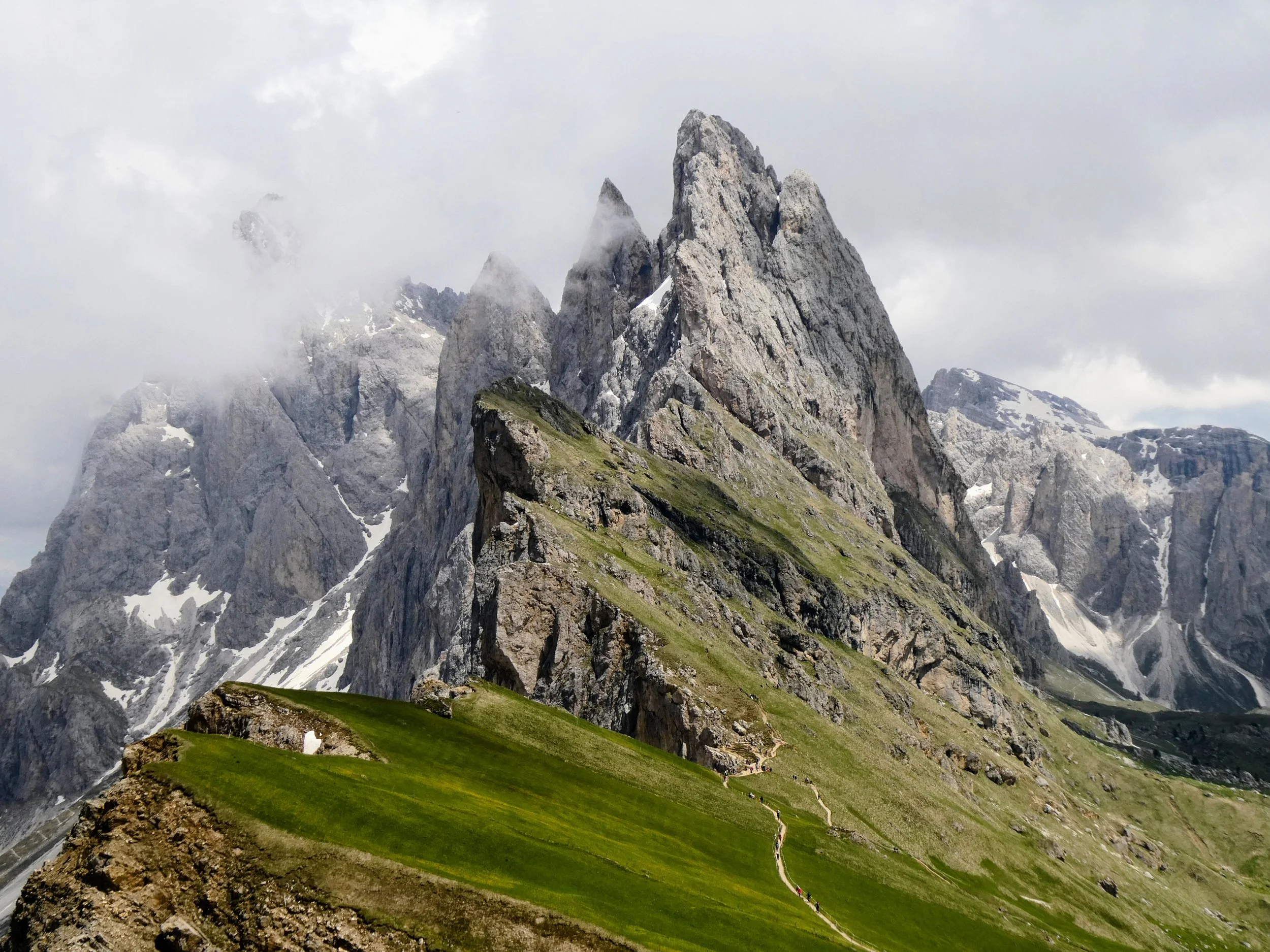 Jagged mountain peaks with snow patches rise above green grassy slopes in a cloudy sky.