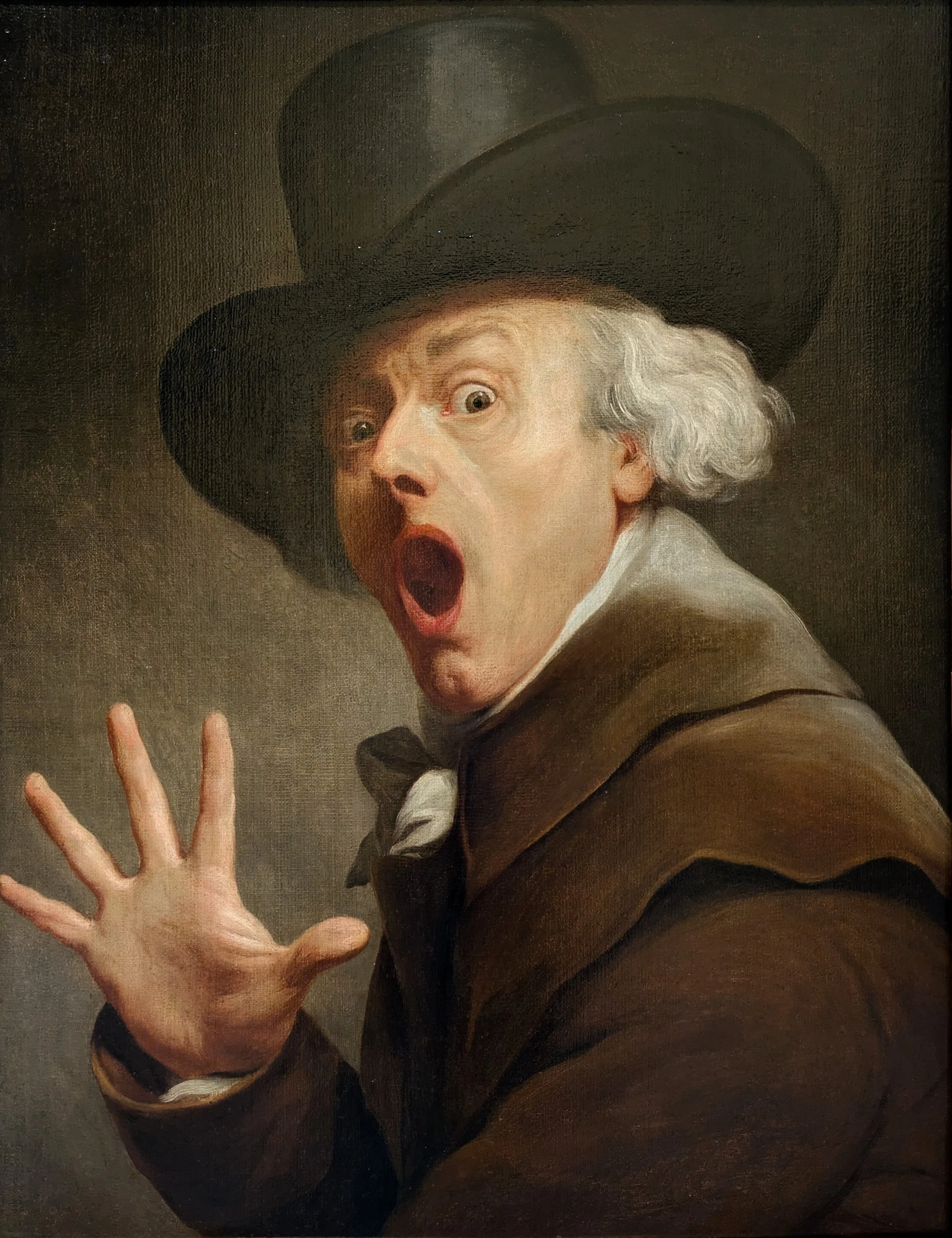 A man with white hair and a surprised expression, wearing a dark hat and brown coat, with his hand raised in front of him.
