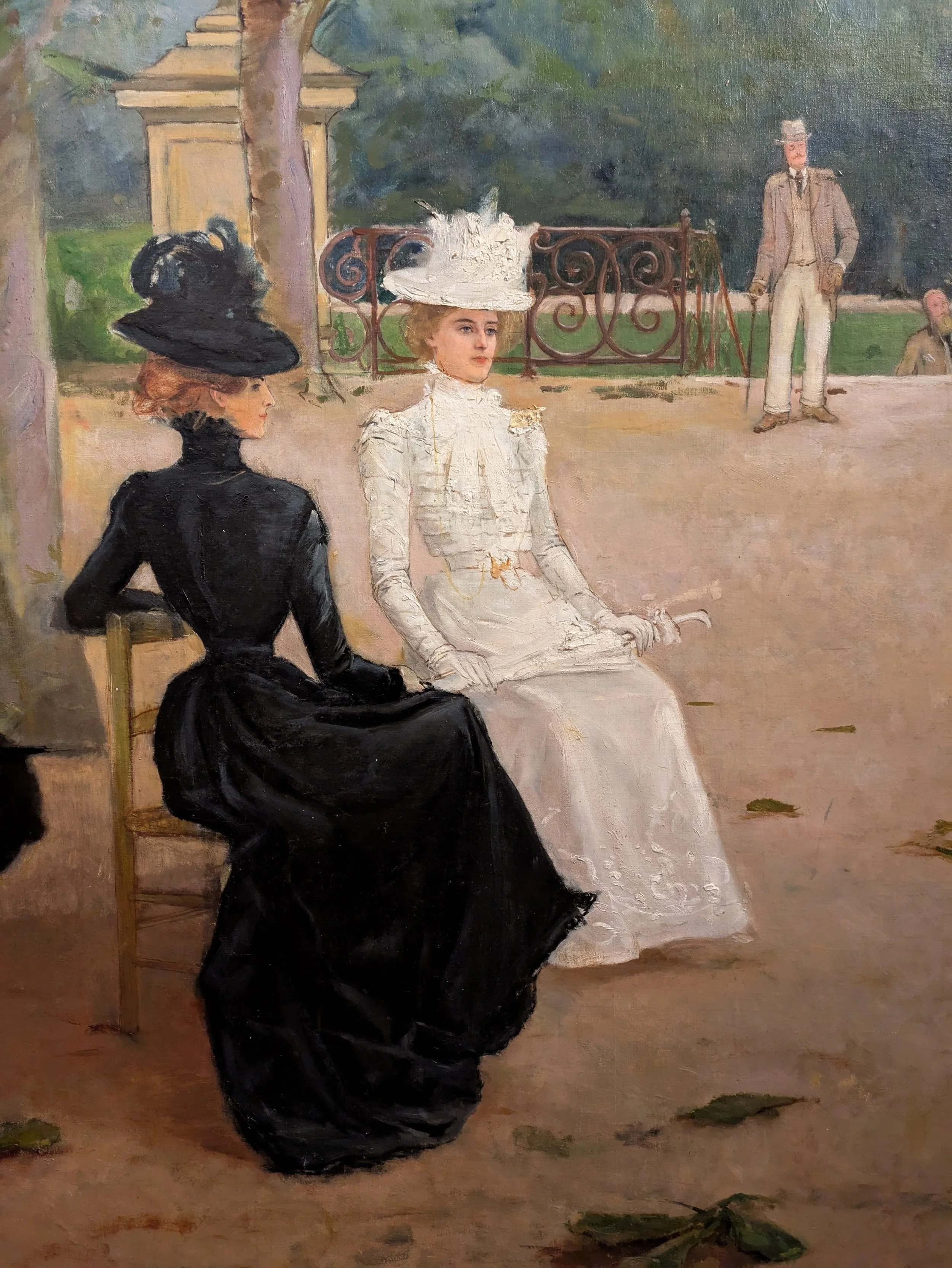 A painting of two women seated outdoors, dressed in early 20th-century fashion. One woman is wearing a black dress and a large black hat, and the other is in a white dress with a wide-brimmed white hat decorated with feathers. In the background, a man in a light-colored suit and hat stands near an ornate black wrought iron fence, with trees and a park-like setting behind them.