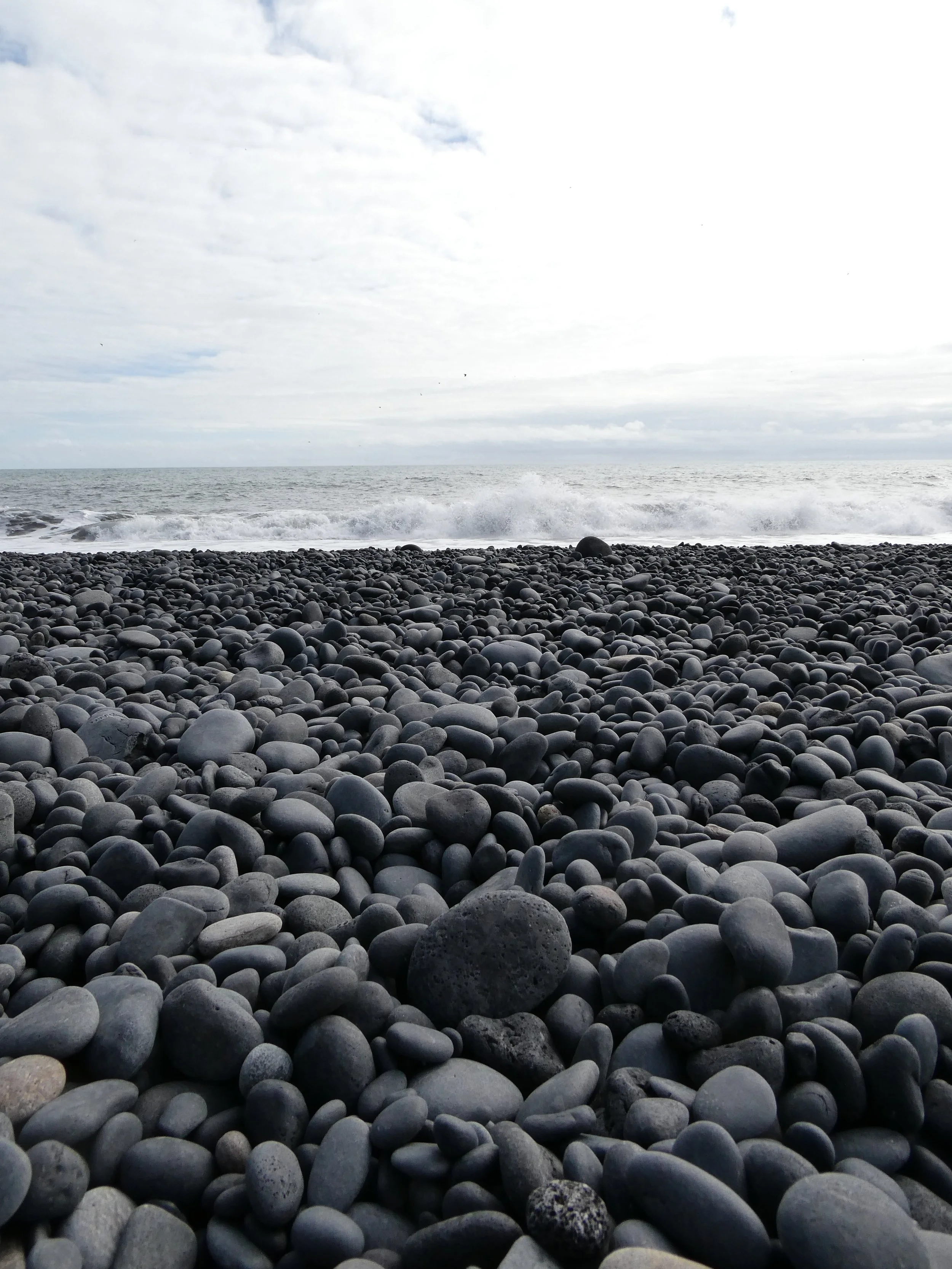 A rocky beach with smooth, rounded stones covering the shore and ocean waves in the background under a cloudy sky.