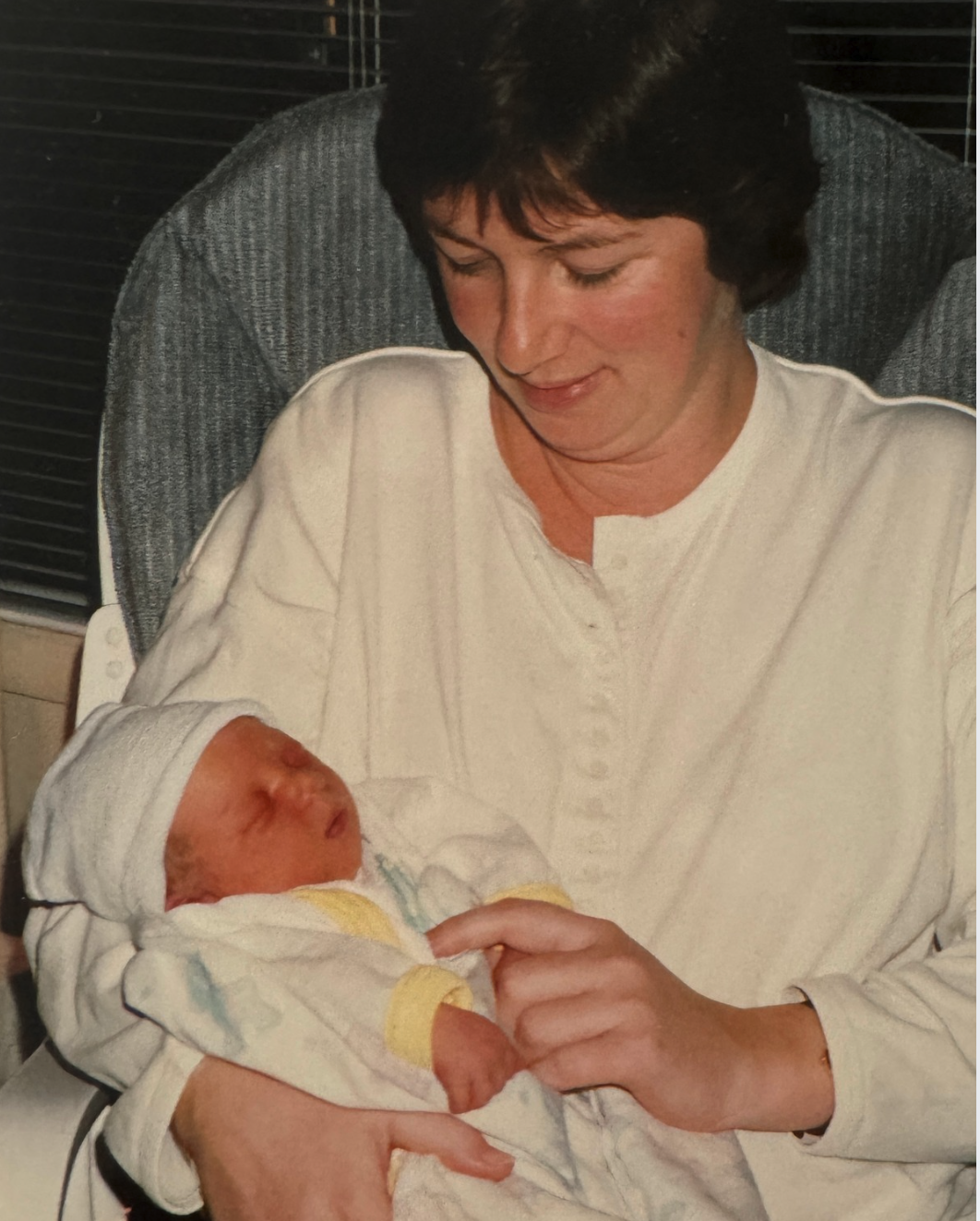 A woman holding a newborn baby in her arms, both dressed in hospital gowns, in what appears to be a hospital room.