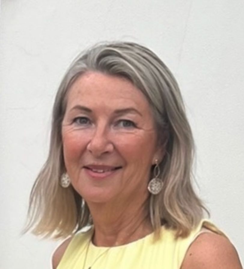A smiling middle-aged woman with shoulder-length blonde hair and earrings, wearing a yellow sleeveless top, standing against a plain light-colored background.