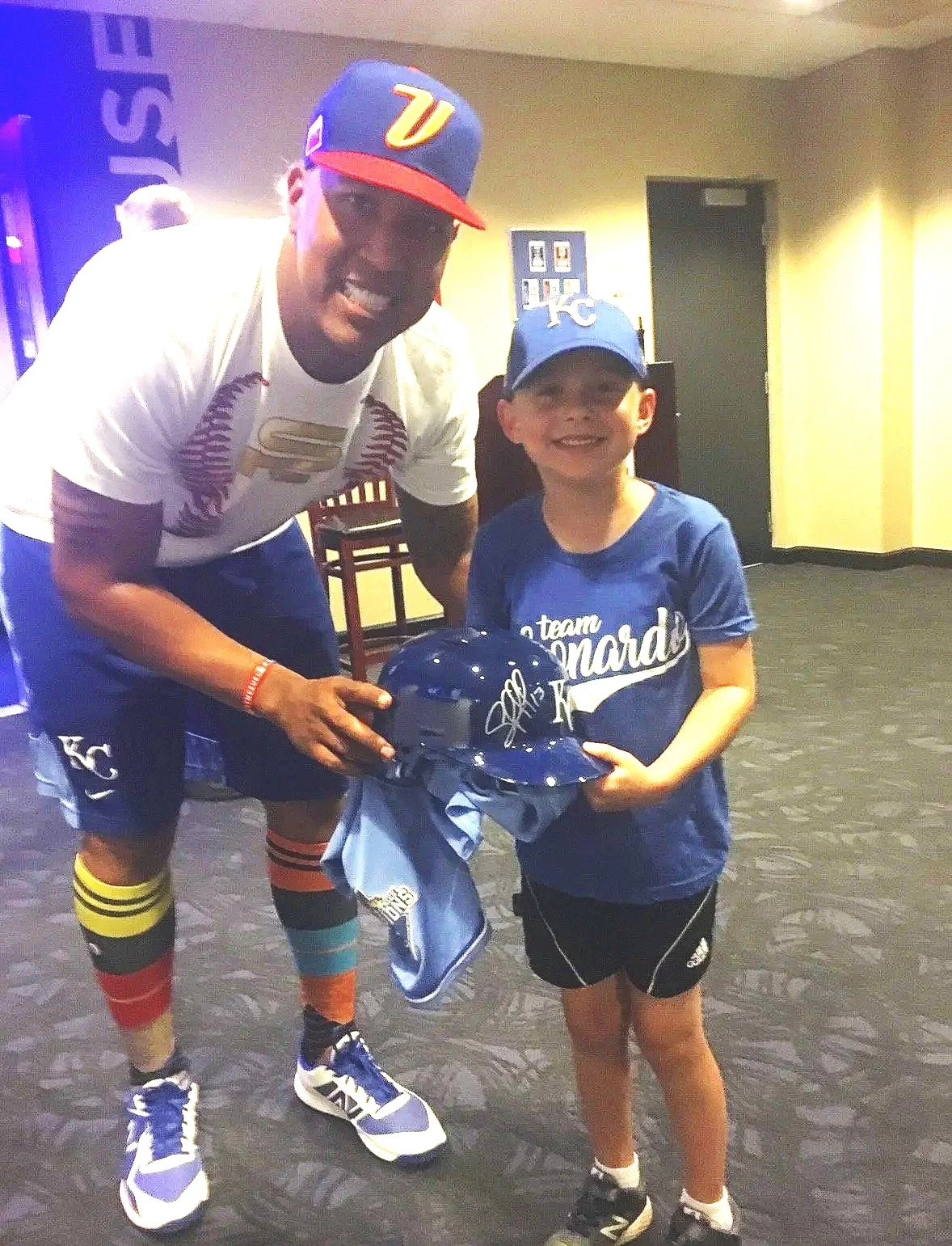 Leonardo and Salvador Perez wearing Kansas City Royals baseball caps are smiling and holding a Royals helmet and jersey in an indoor setting.