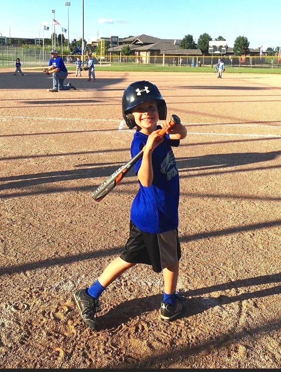 Leonardo smiling in baseball gear holding a bat on a baseball field during daylight, with other players practicing in the background.