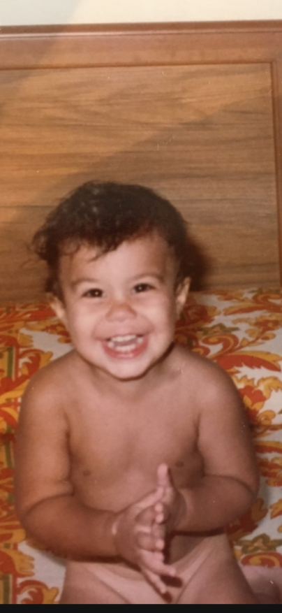 Dominic with curly hair smiling and clapping while sitting on a bed with a colorful patterned blanket.