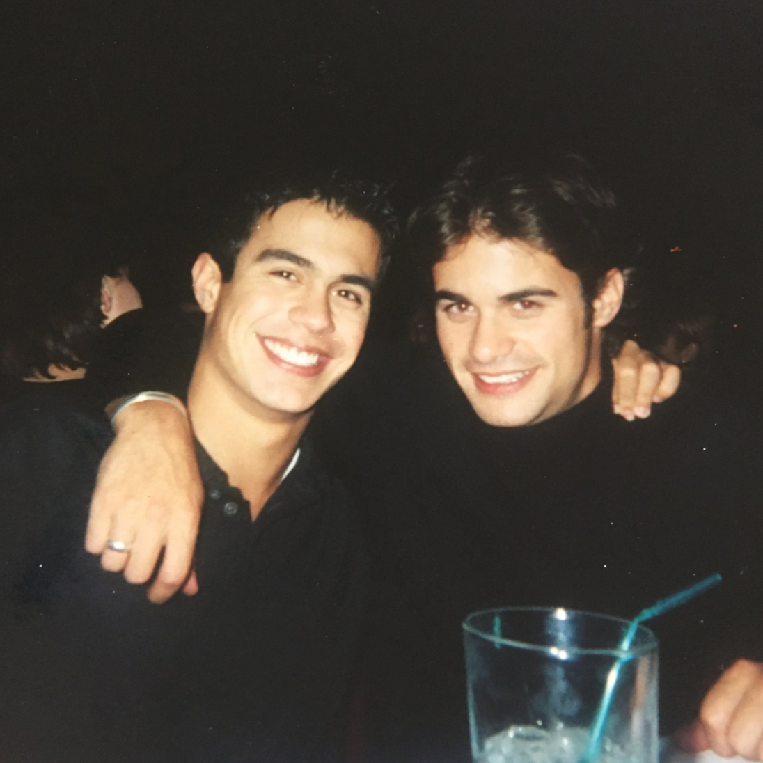 Dante and Dominic smiling and posing with their arms around each other at a social gathering, with a glass and straw visible on the table in front of them.