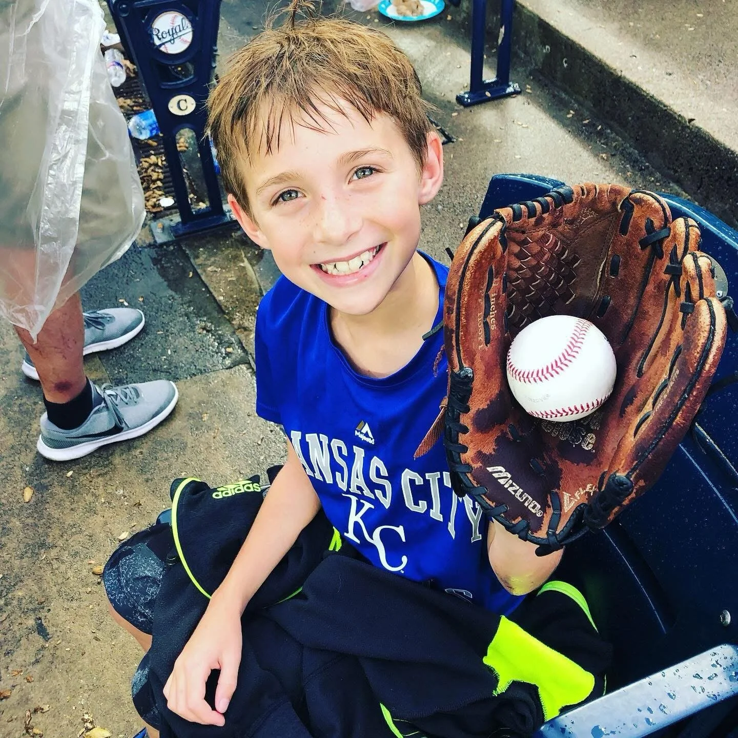 A young boy with wet hair and a big smile, wearing a blue Kansas City Royals T-shirt, sitting on a bench. He's holding a baseball glove with a baseball inside. There are people standing nearby, and the setting looks like a baseball park or dugout with concrete and metal surroundings.