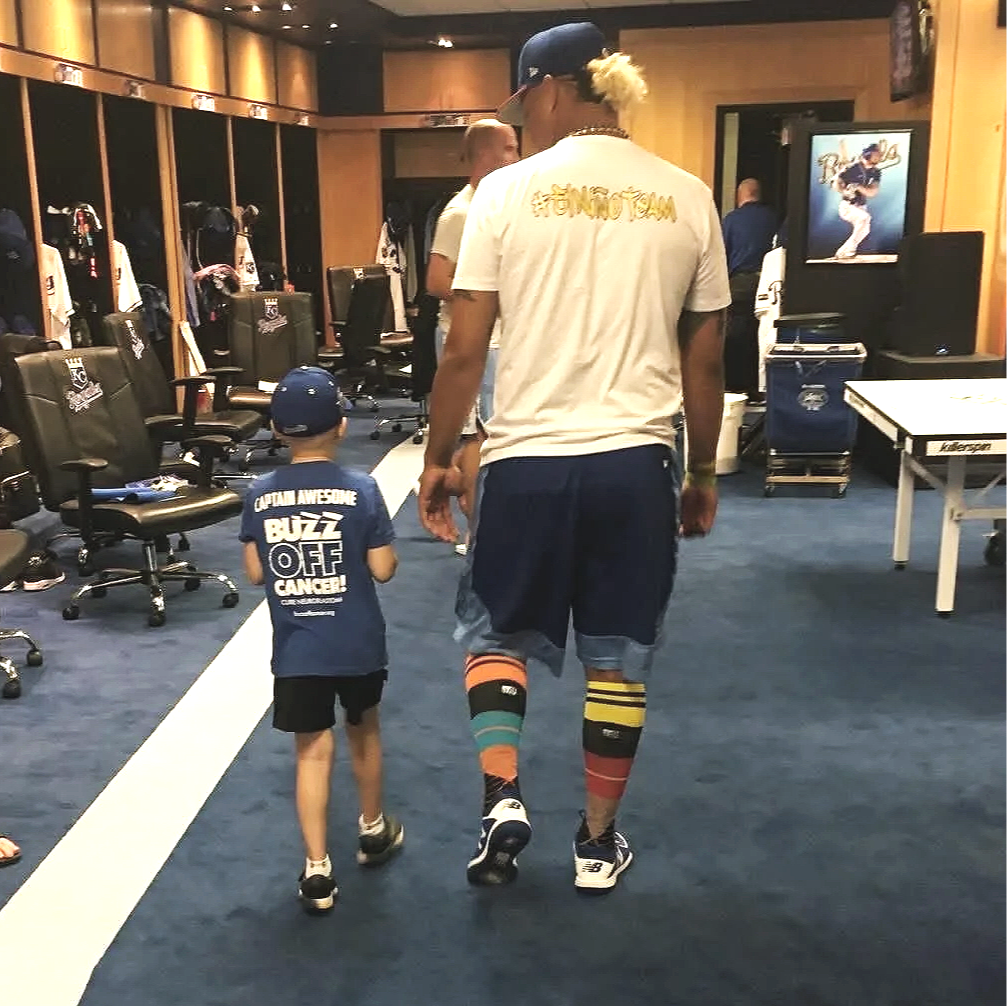 A man and a young boy walking together in a sports locker room. The man is wearing colorful knee-high socks, a baseball cap, and casual athletic clothing. The boy is dressed in a baseball uniform and a blue cap, with the back of his shirt reading 'Captain Awesome Buzz Off Cancer!'.