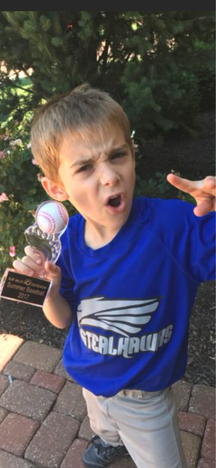 Leonardo holding a baseball trophy in one hand and making a gesture with the other while outdoors. He has short, light brown hair, and wearing a blue shirt with a winged logo and beige pants.