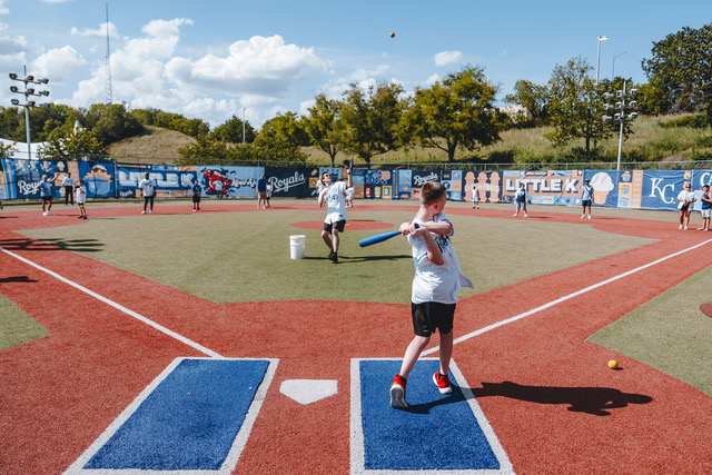 Children practice baseball at a local sports field on a sunny day, with trees and a blue sky in the background.