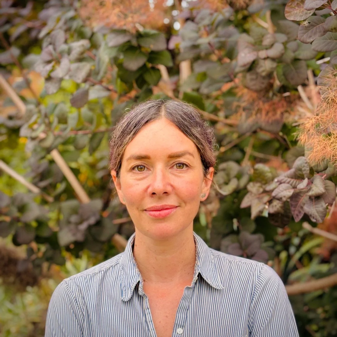 An attentive woman with brown hair, middle-parted and pulled back, and fair golden skin stands outdoors in front of a lush, leafy background with purple and green foliage, wearing a plain striped button-up shirt.