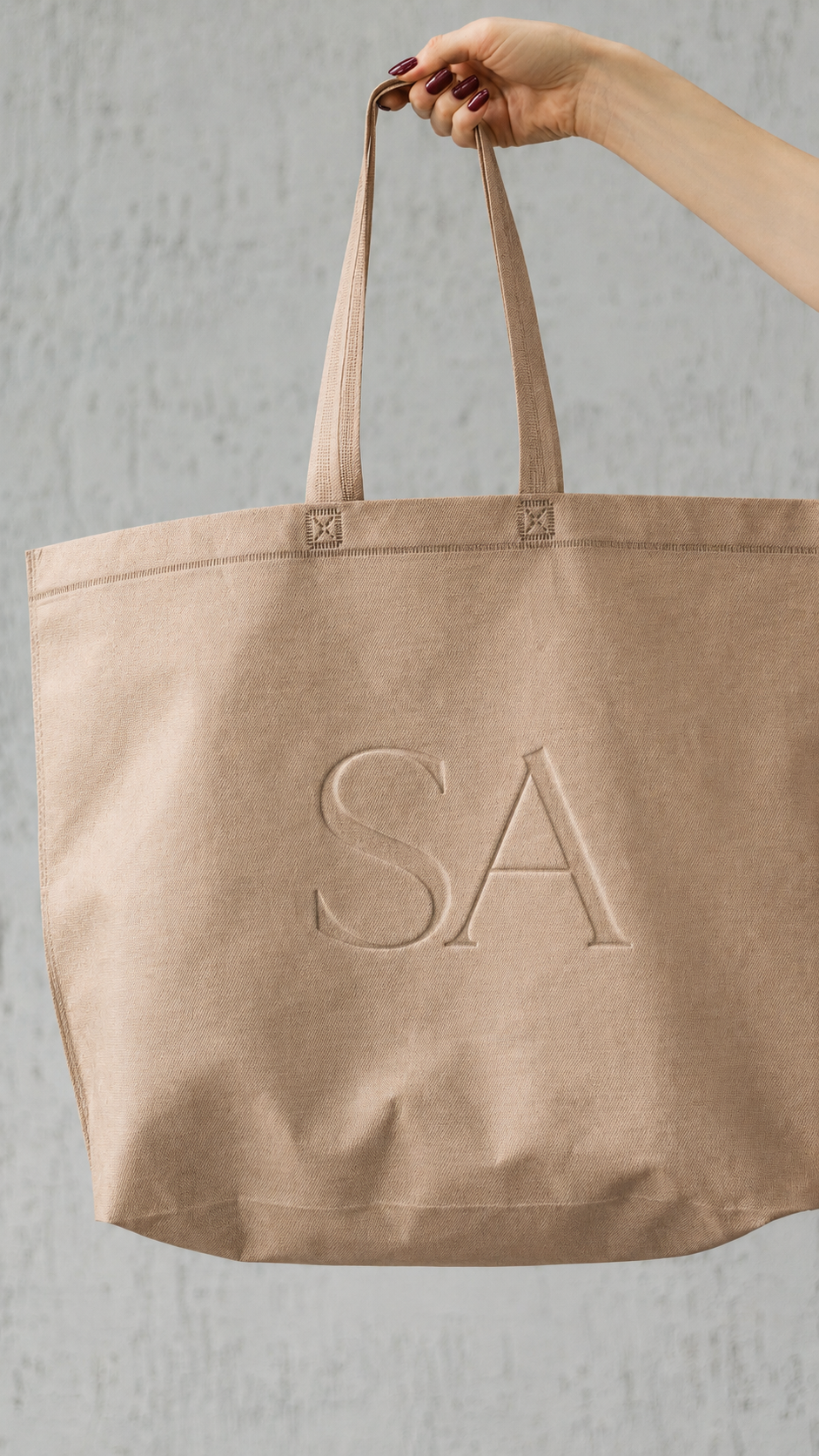 A beige canvas tote bag with the letters 'SA' embossed on the front, held by a hand with dark red nails against a gray textured background.