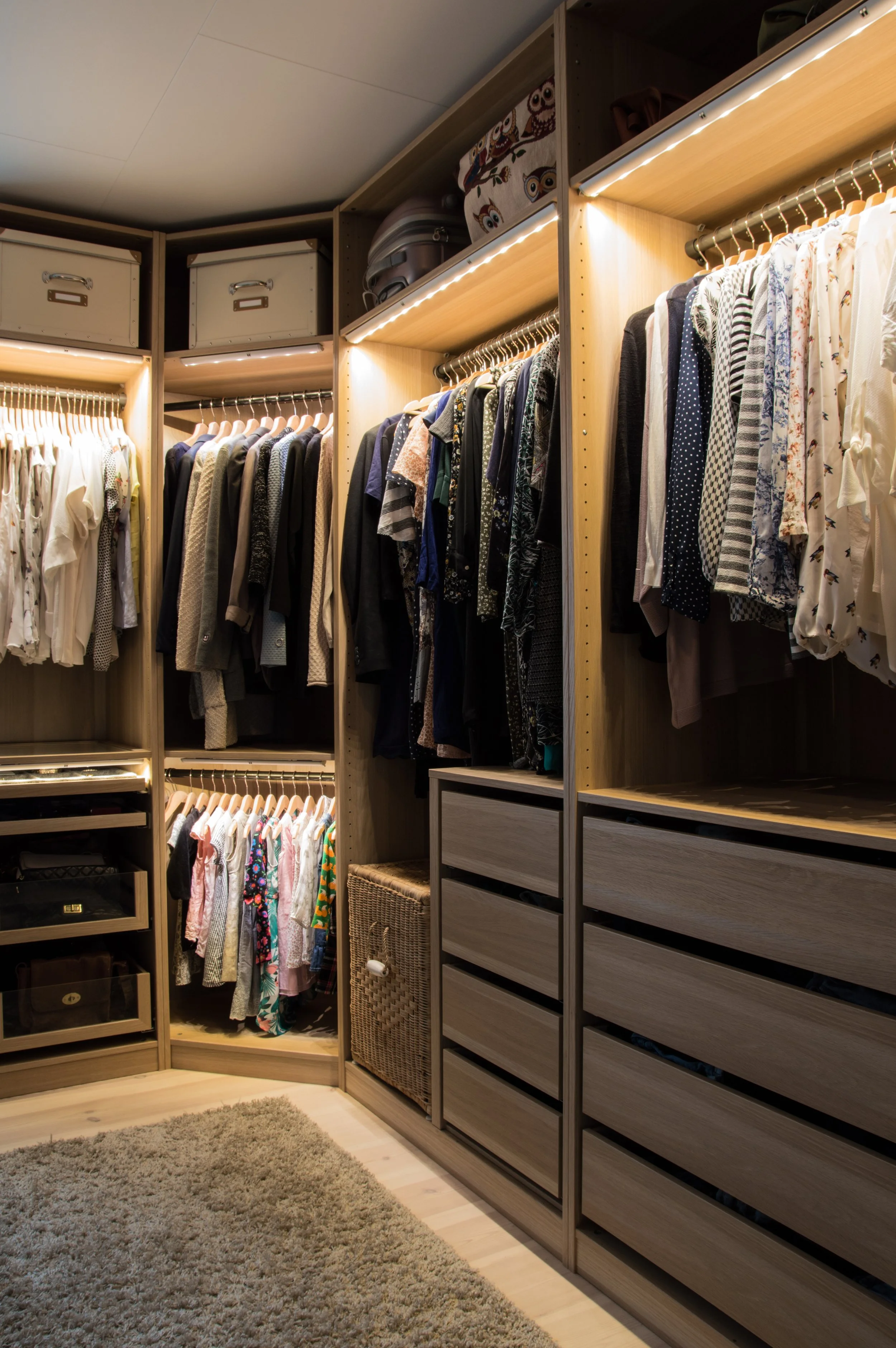A walk-in closet with wooden shelves and hanging clothes, including shirts, dresses, and jackets. There are storage boxes on the top shelves and a wicker basket. The floor is wooden with a beige rug in front of the shelves.