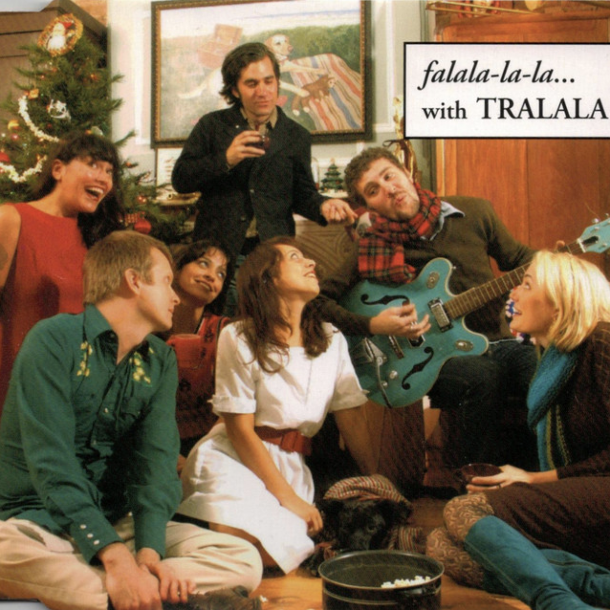 The album cover of Falalalala with Tralala features seven people celebrating the winter holidays around a decorated Christmas tree, some sitting on the floor, others standing. They are smiling and talking, with a festive, cozy indoor setting.