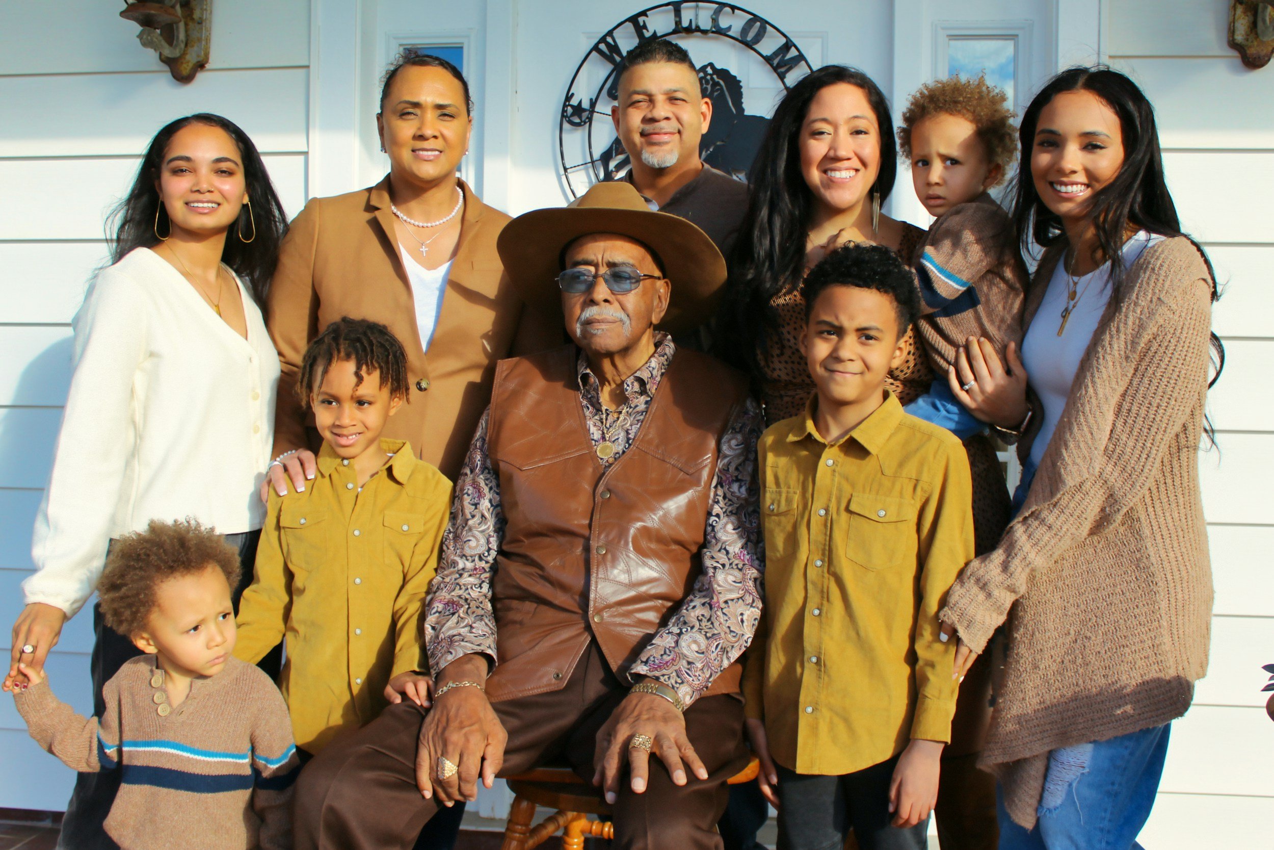 A diverse multigeneration family gathered outdoors in front of a house with a "Welcome" sign. They are smiling and posing for the photo, with a mix of adults and children, some standing and one older man seated in front.