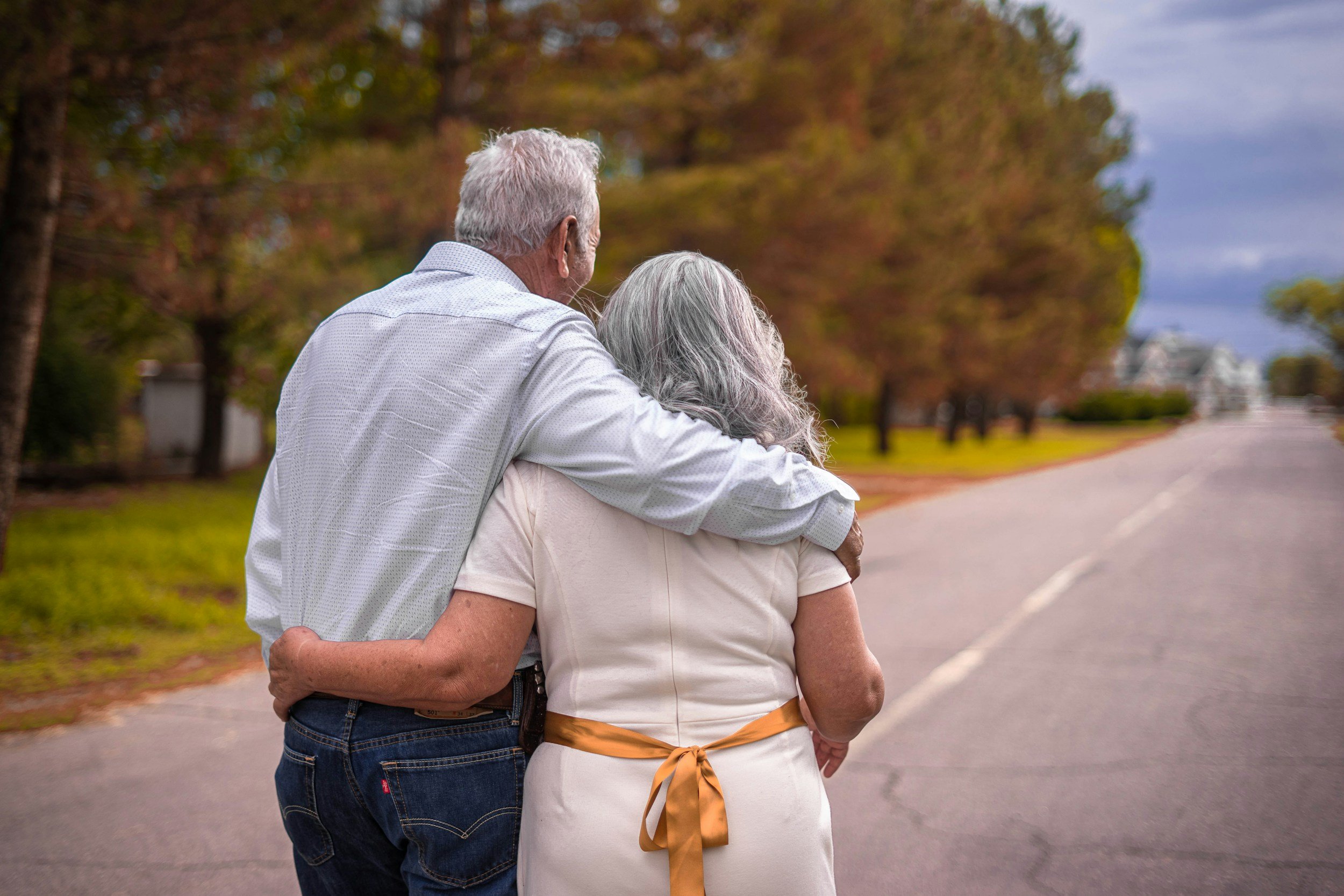 An elderly couple walking outdoors on a sidewalk, with the man resting his arm on the woman's shoulder, surrounded by trees with autumn foliage.