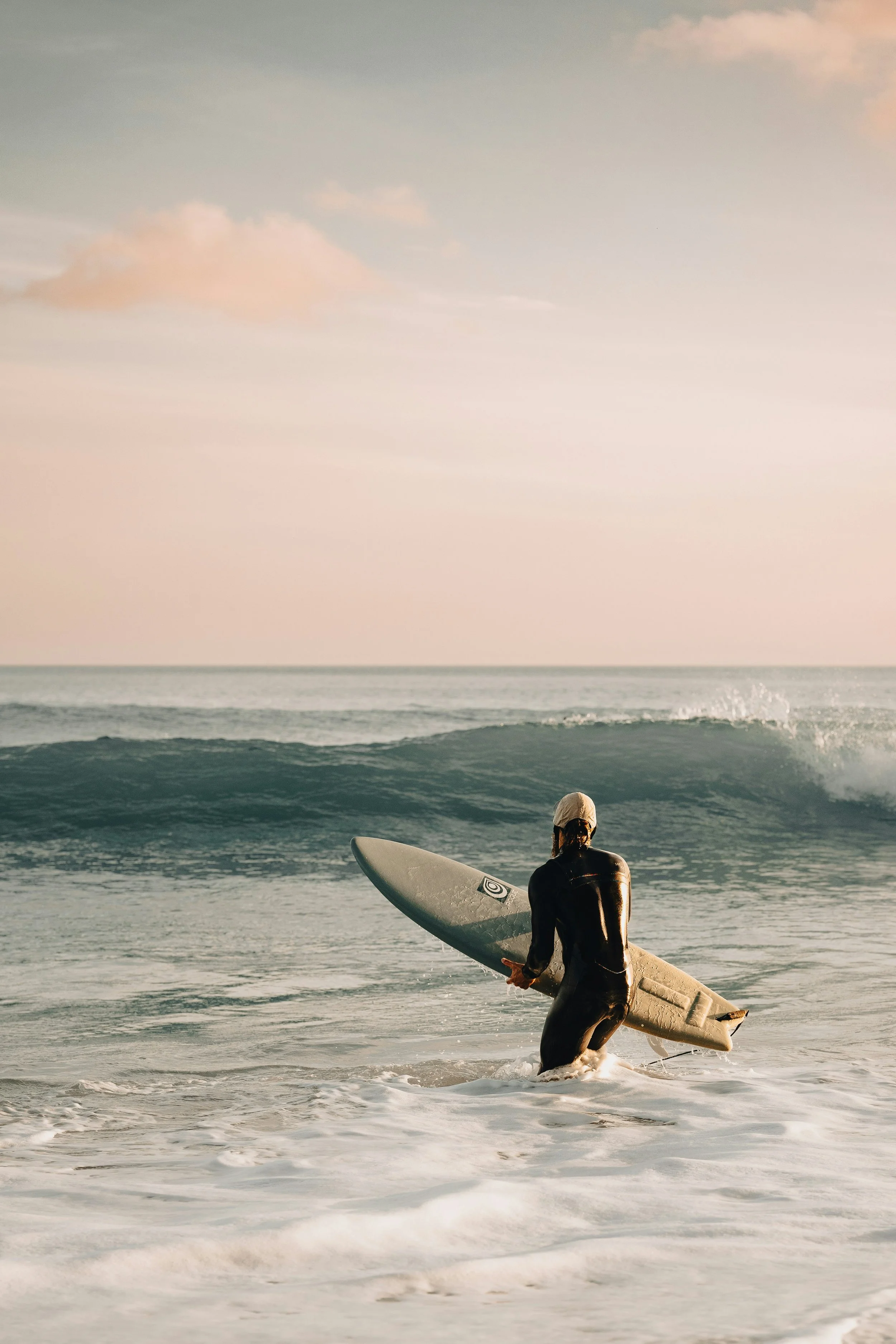 A surfer in a wet suit carrying a surfboard into the ocean at sunset, with waves and a cloudy sky in the background.