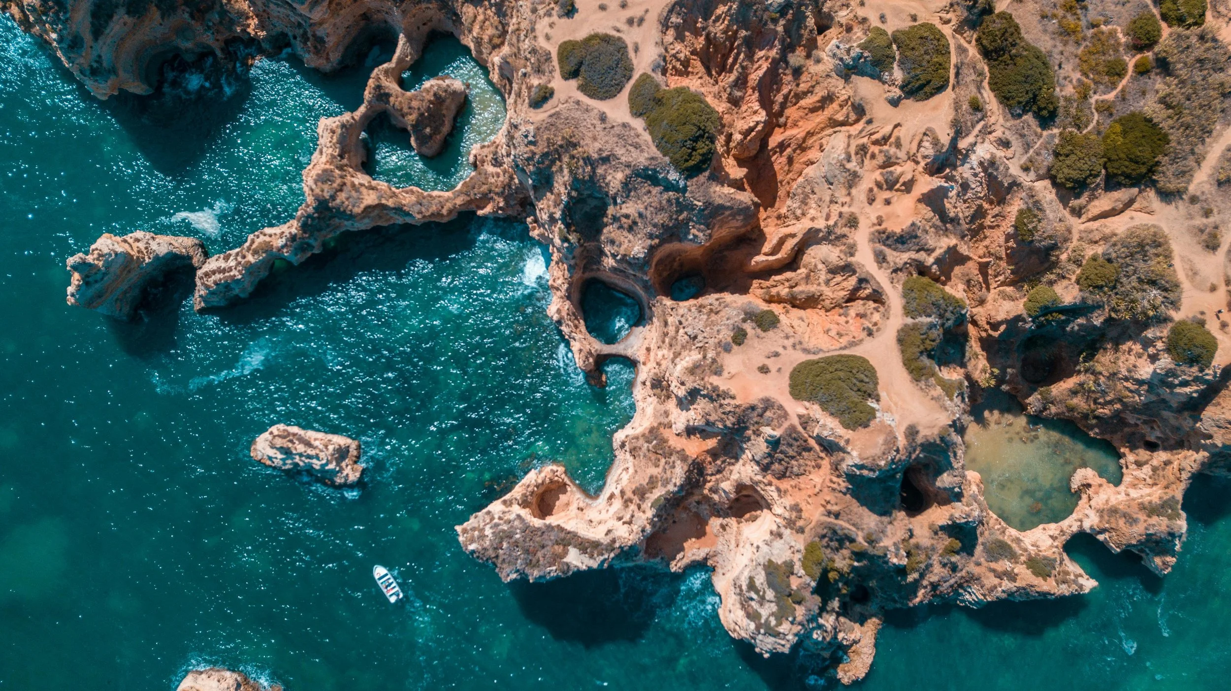 Aerial view of a rocky coastal area with turquoise water, featuring small islands and caves, with a boat swimming nearby.