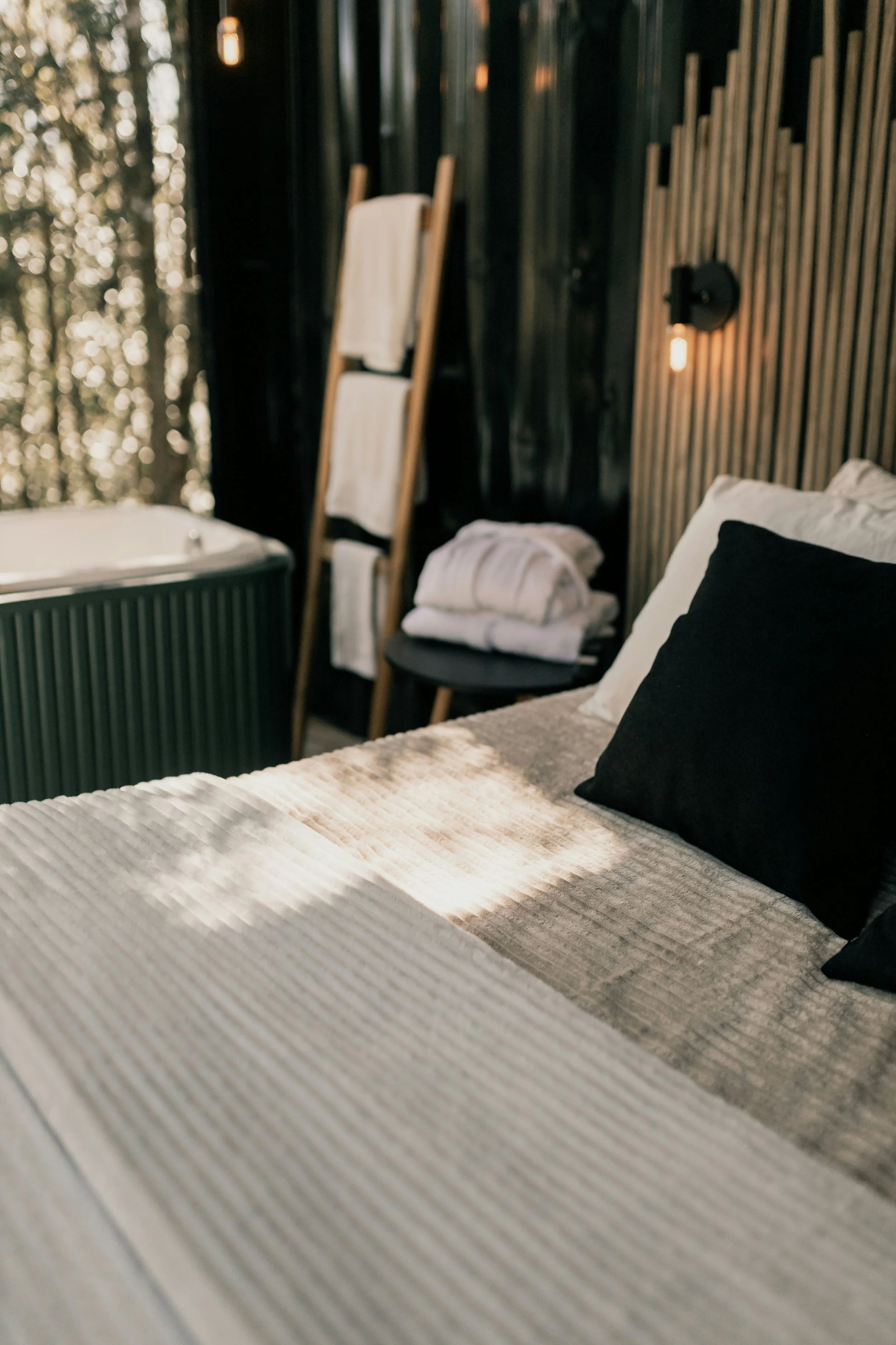 A cozy bedroom with a bed, black and white pillows, a beige textured blanket, a black side table with folded towels, a chair with towels, a wooden ladder, and a hot tub, with dark wood paneling on the walls and warm lighting. Outside, there are trees visible through a window.