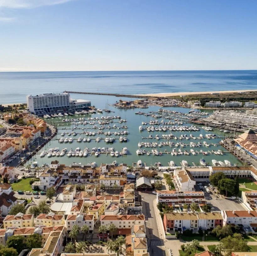 Aerial view of a marina with boats, surrounded by residential buildings and a beach with the ocean in the background.