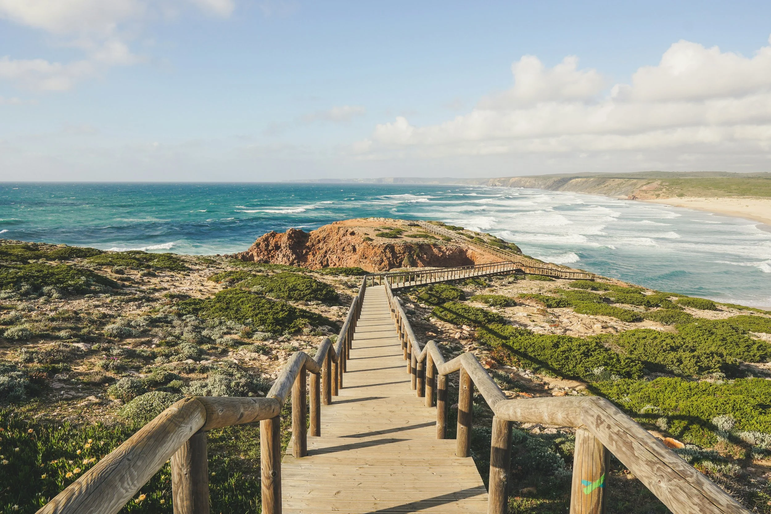 A wooden pathway leading to a beach with waves and cliffs in the distance under a partly cloudy sky.