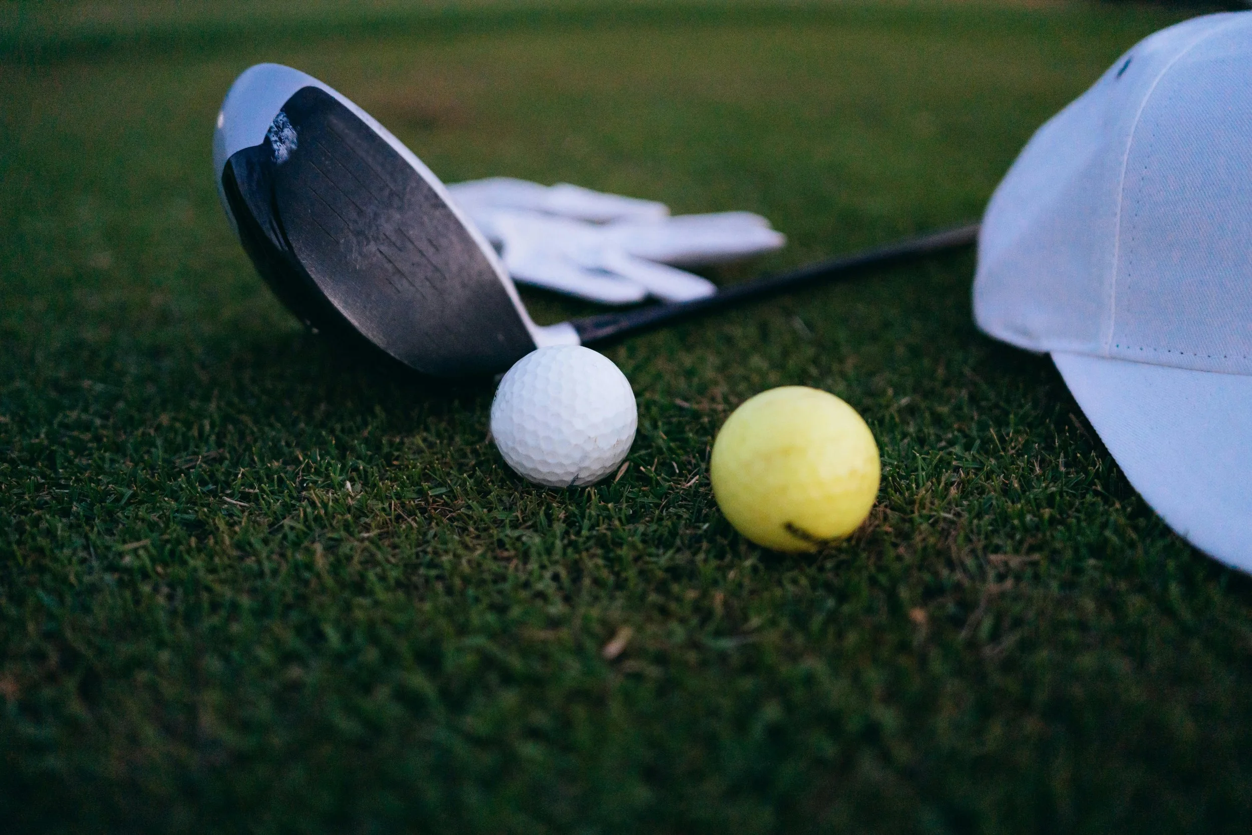 Close-up of two golf balls, one white and one yellow, on a grassy surface with a golf club and a white golf cap nearby.