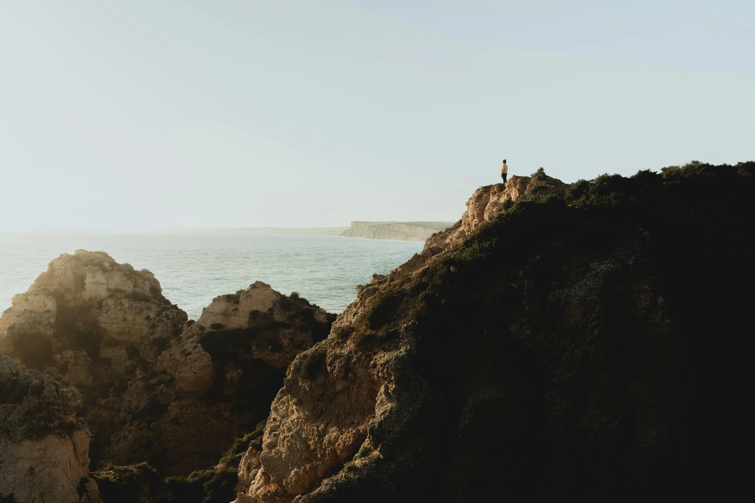 A woman standing on a rocky cliff overlooking the ocean with cliffs in the distance.