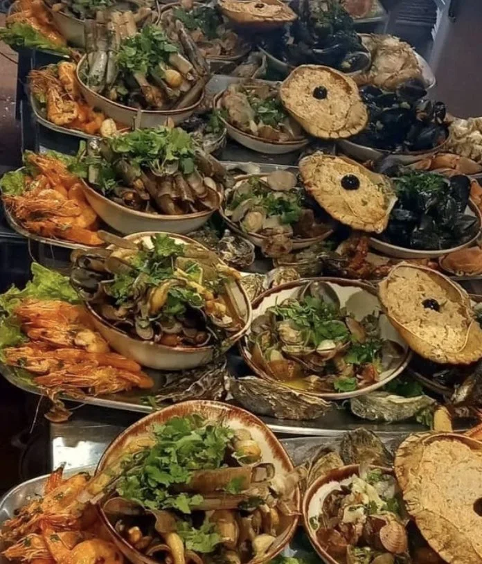 A table filled with various seafood dishes, including shells, shrimp, and oysters, garnished with herbs and lemon slices.