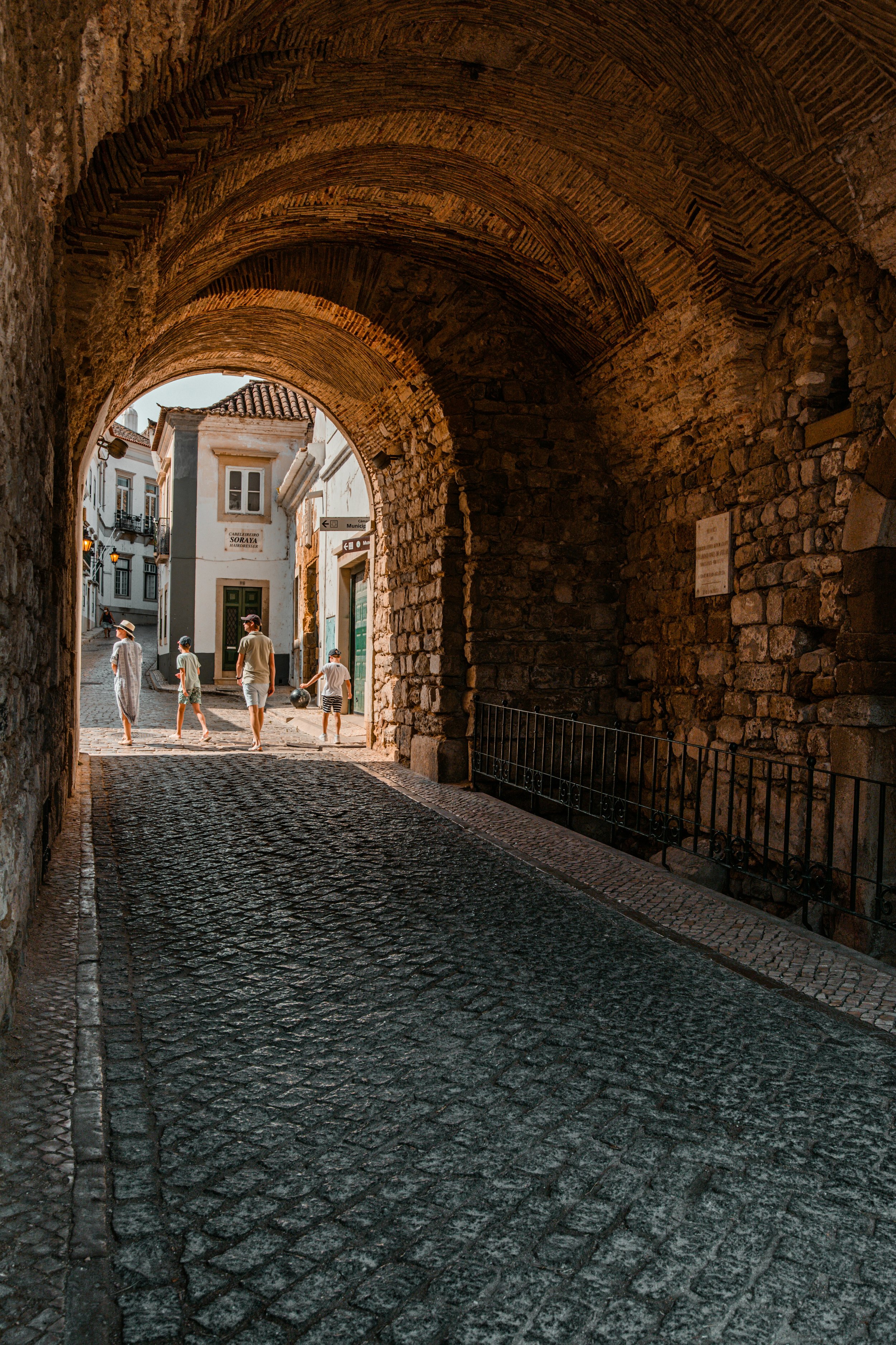 A cobblestone street framed by an ancient brick archway, leading to a sunny square with people walking and historic white buildings.
