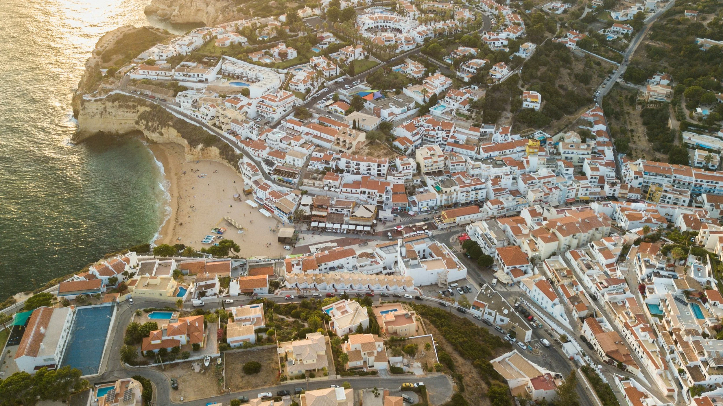 Aerial view of a coastal town with white buildings and red-tiled roofs, a sandy beach, and the ocean. The area includes residential and commercial structures, roads, and greenery.