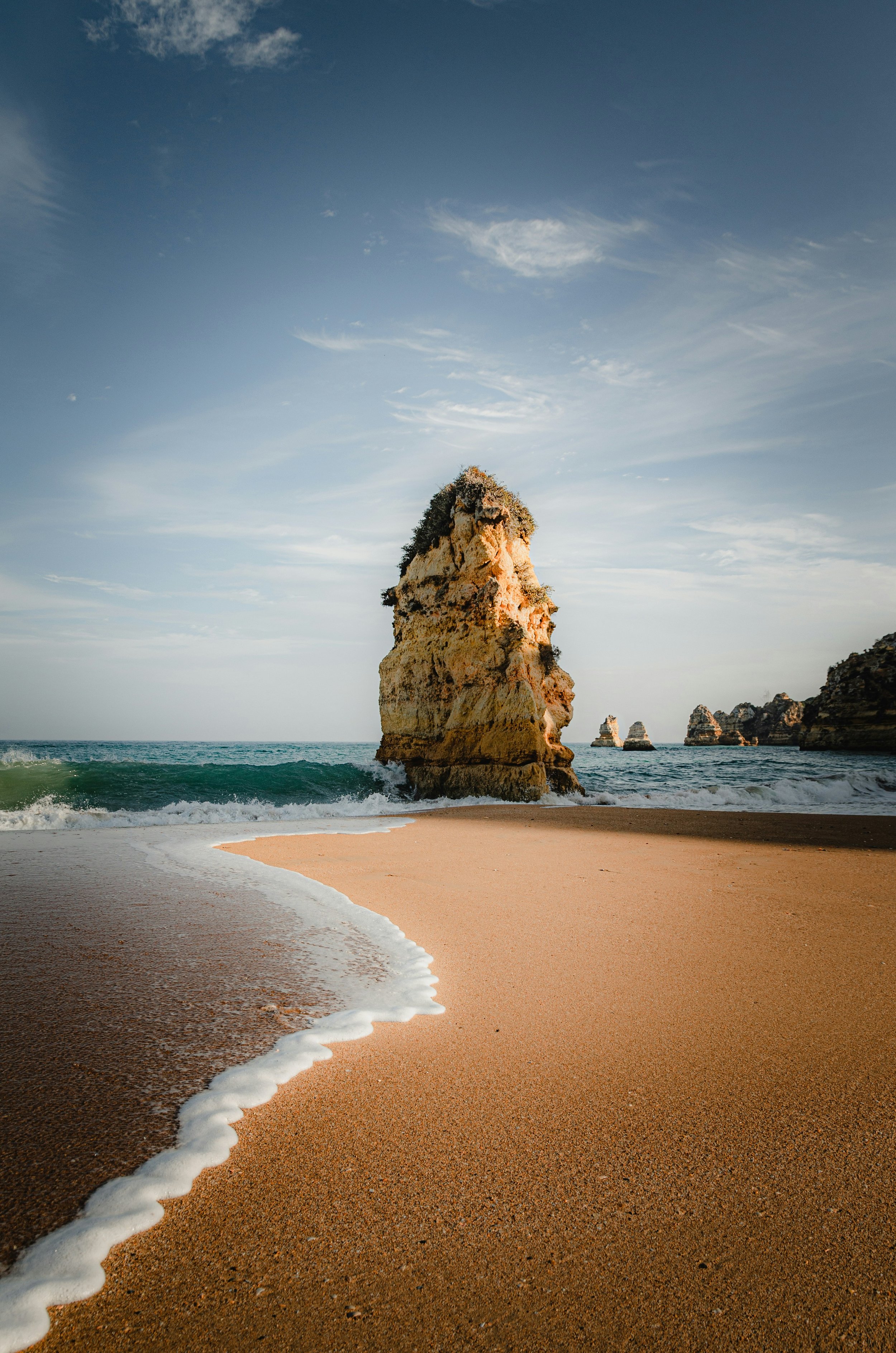 Beach scene with three large rock formations in the ocean and a sandy shore in the foreground, under a partly cloudy sky.