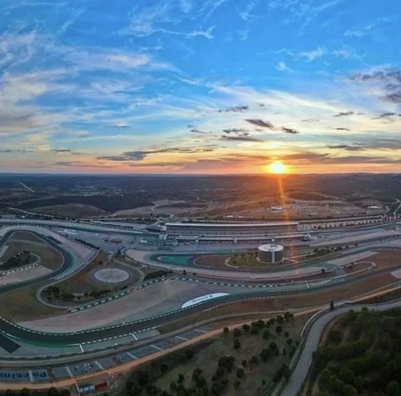Aerial view of a racetrack complex with curves and straightaways, surrounded by roads and greenery, capturing a sunset in the sky with clouds and sunlight casting a warm glow.
