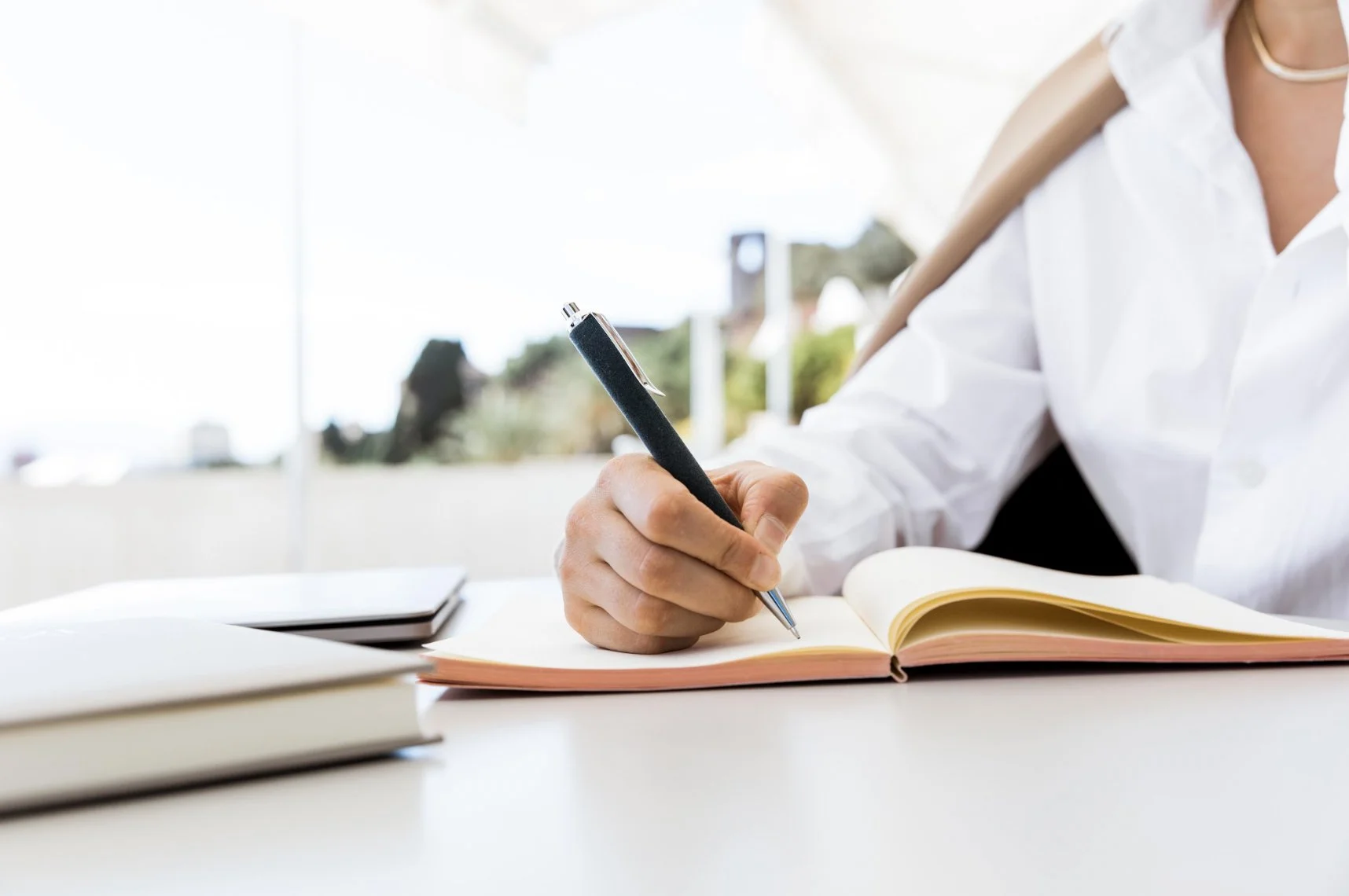 A person in a white shirt writing in a notebook with a pen, on a desk with books and a laptop, with a blurred outdoor background.
