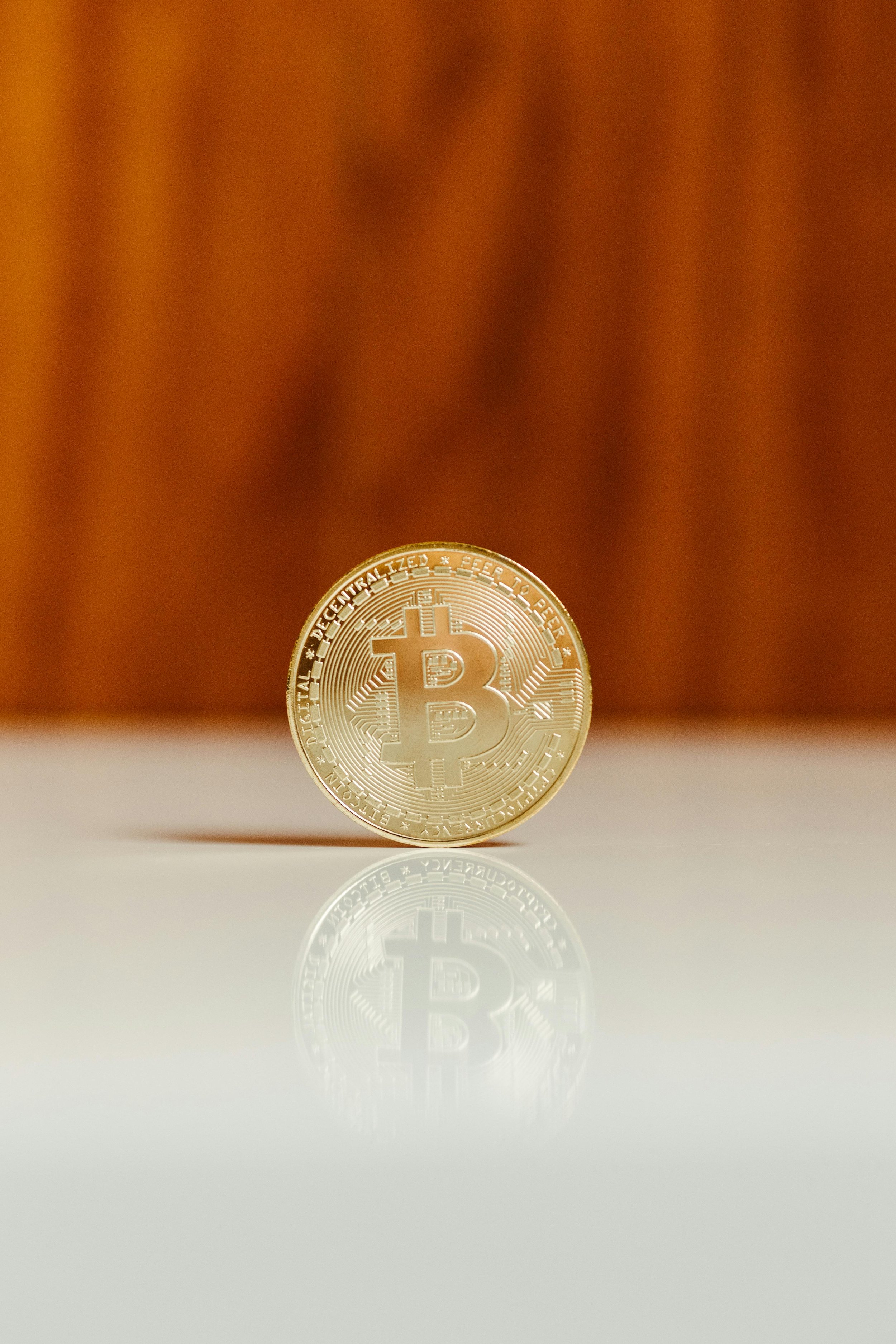 A gold-colored Bitcoin coin with the Bitcoin symbol in the center, placed on a reflective surface, with a brownish background.