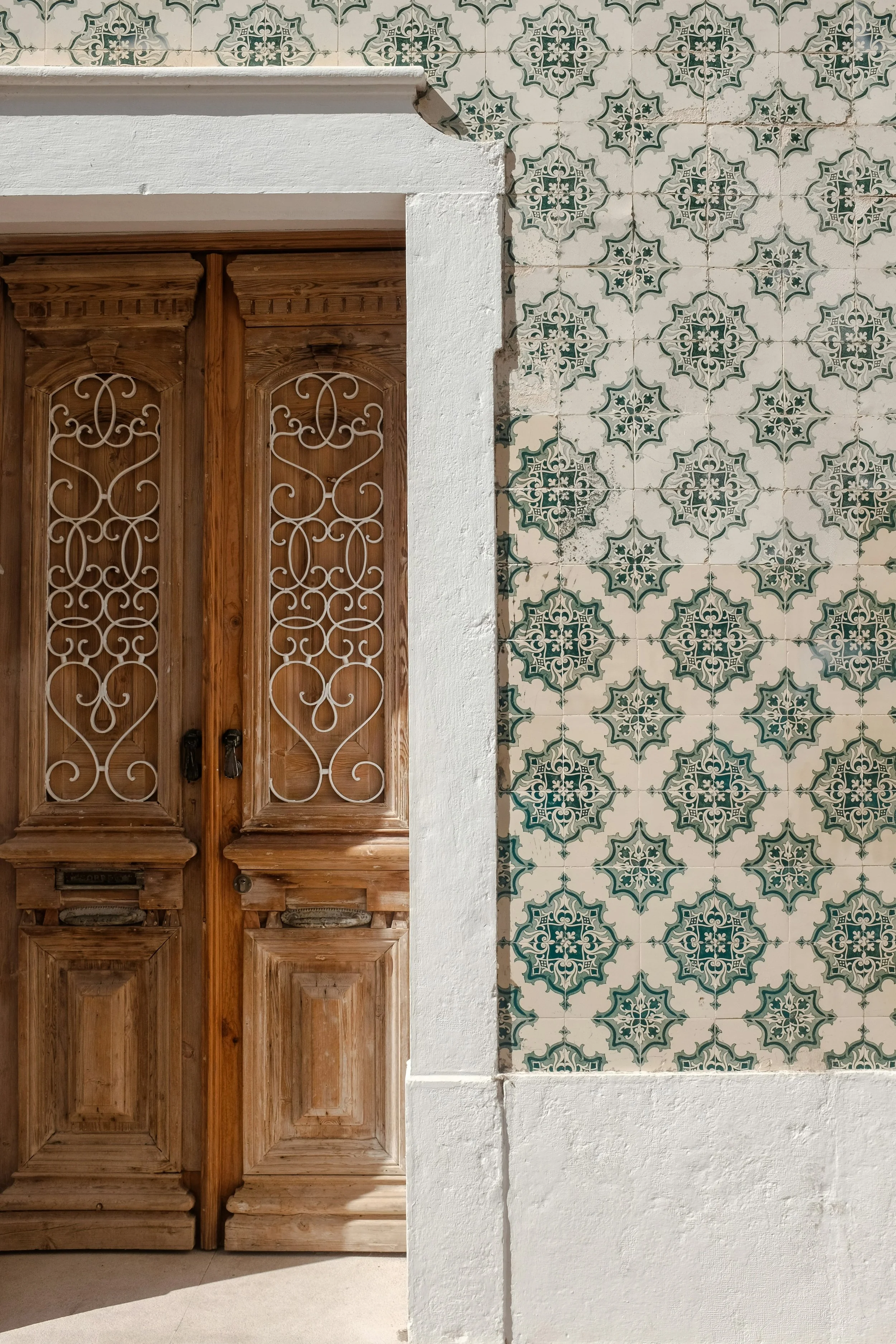 Wooden double doors with decorative wrought iron grillwork, set in a white wall with traditional patterned ceramic tiles on the right side.