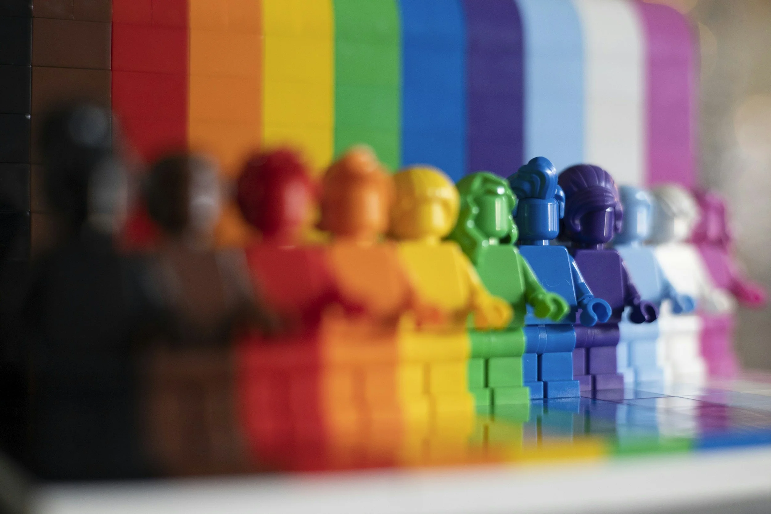A row of colorful LEGO figurines in various colors including black, red, orange, yellow, green, blue, purple, and pink, standing in front of a large rainbow-colored building blocks background.