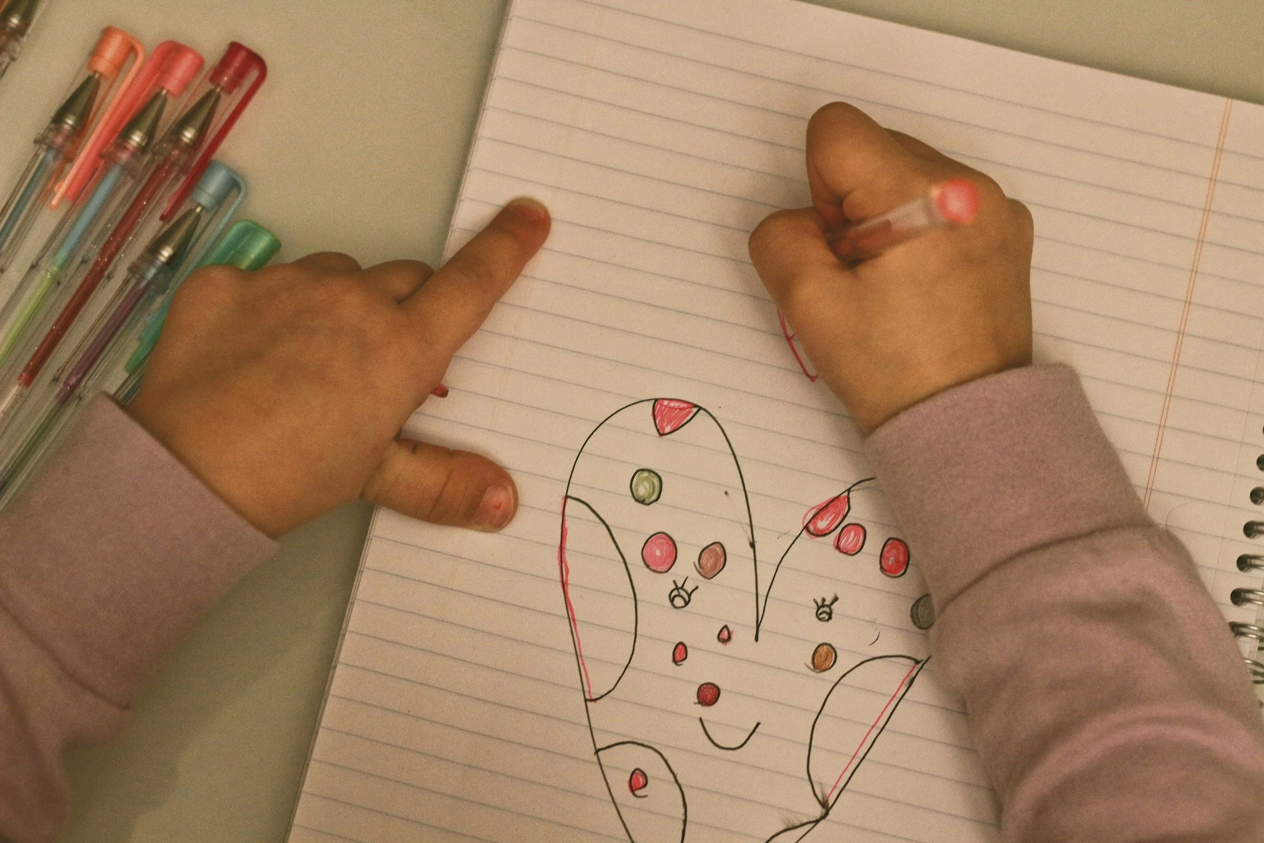 A child is drawing on a lined notebook paper with a pink crayon. The drawing appears to be a colorful ladybug with red, black, and green spots. There are multiple gel pens with pastel-colored caps placed on the desk nearby.