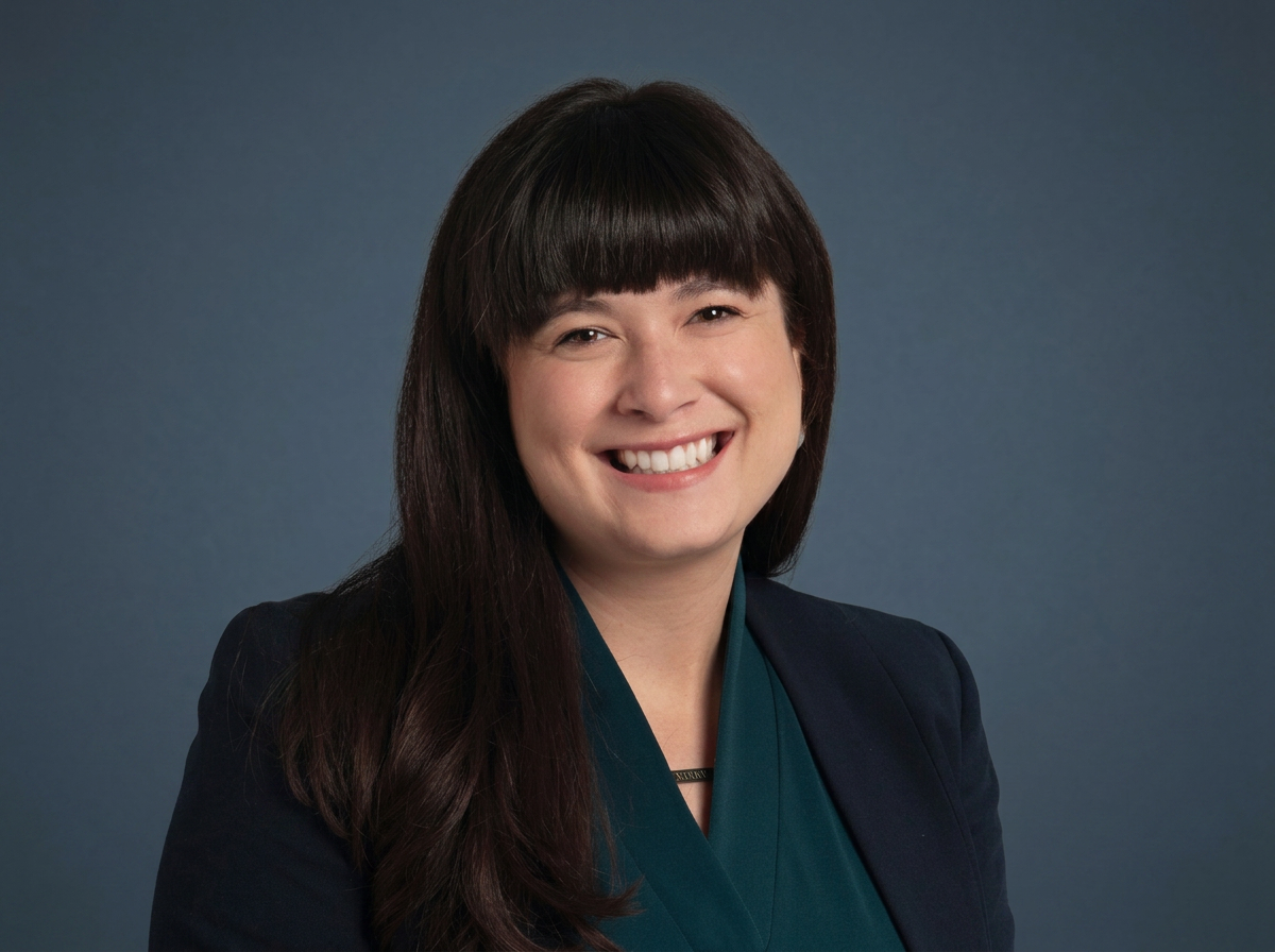 A woman with long dark hair and bangs wearing a dark blazer and teal blouse, smiling against a gray gradient background.