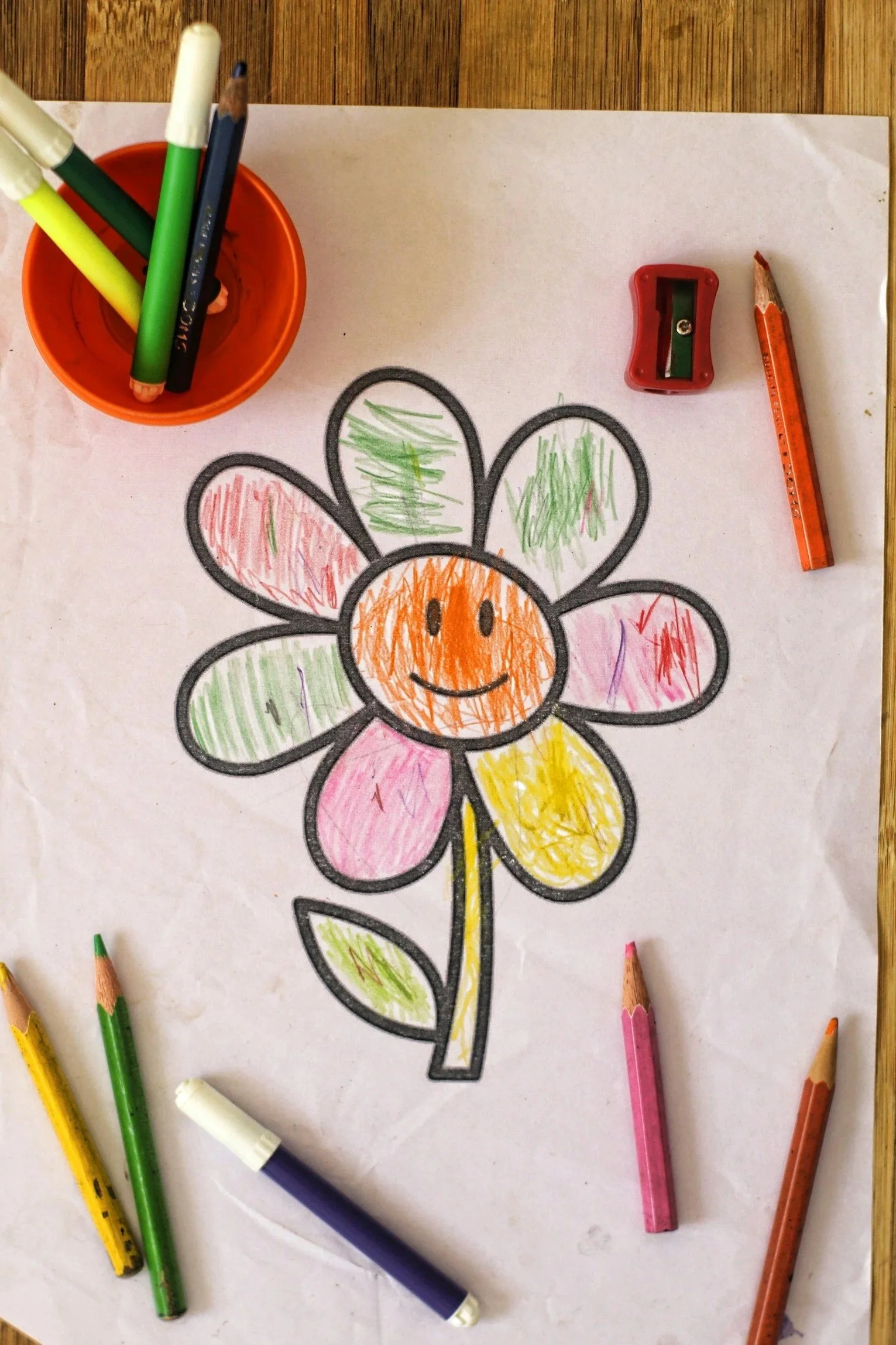 Colorful drawing of a smiling flower with multicolored petals on a white sheet of paper, surrounded by various colored markers and a small red pencil sharpener.