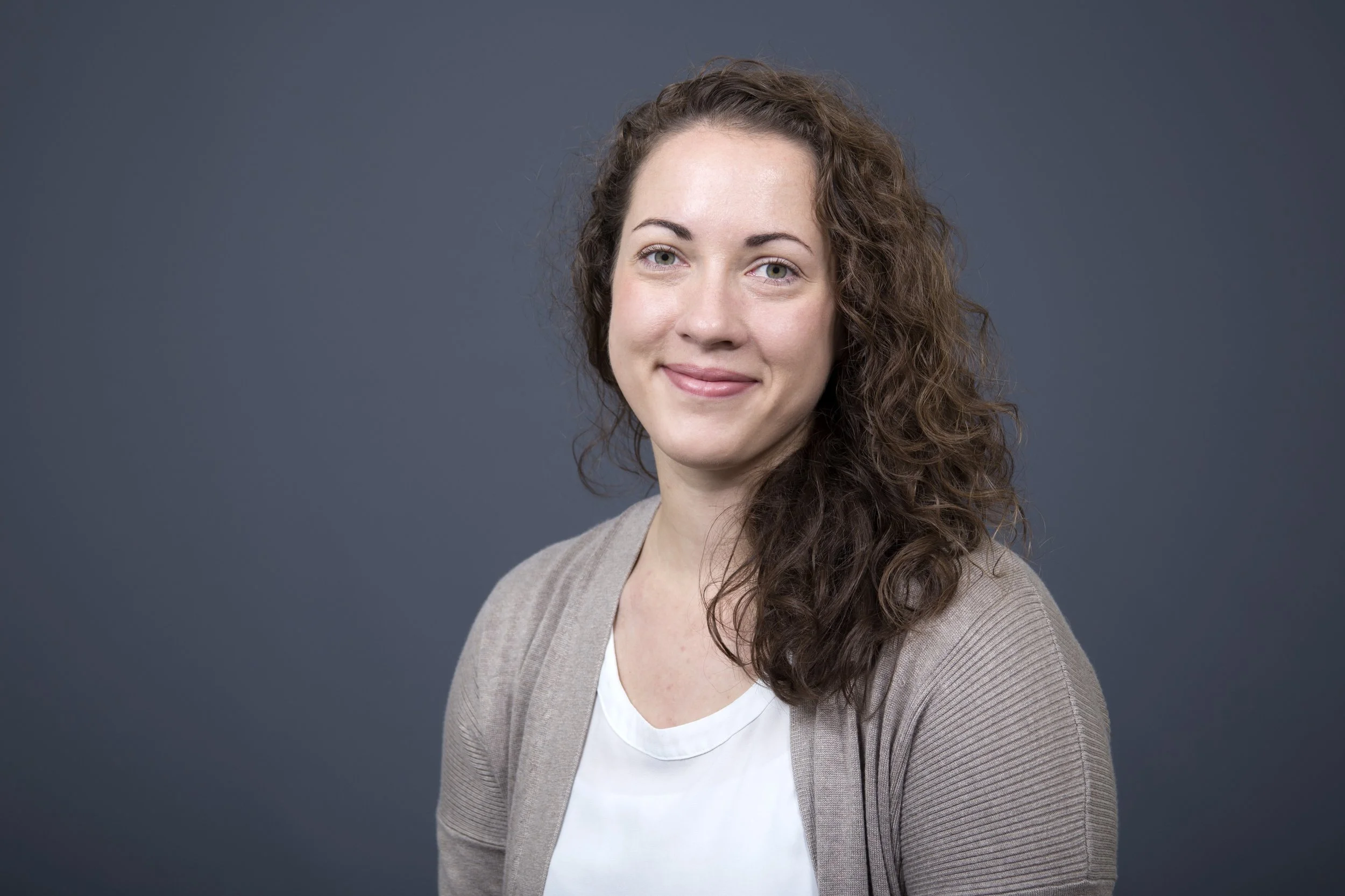 Portrait of a woman with curly brown hair, wearing a beige cardigan and white top, smiling against a plain dark grey background.