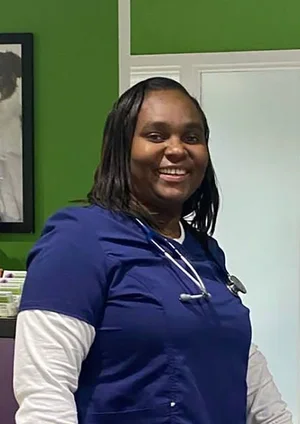 A smiling woman in blue nursing scrubs with a stethoscope, standing in a room with green walls and a whiteboard.