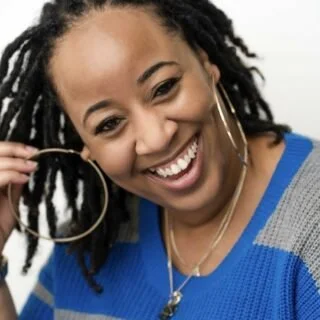 A woman with curly hair wearing large hoop earrings and a blue sweater, smiling and looking at the camera.