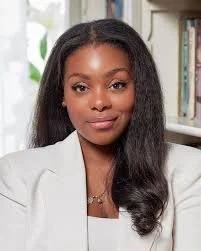 A woman with long black hair wearing a white blazer, standing in front of a bookshelf filled with books.