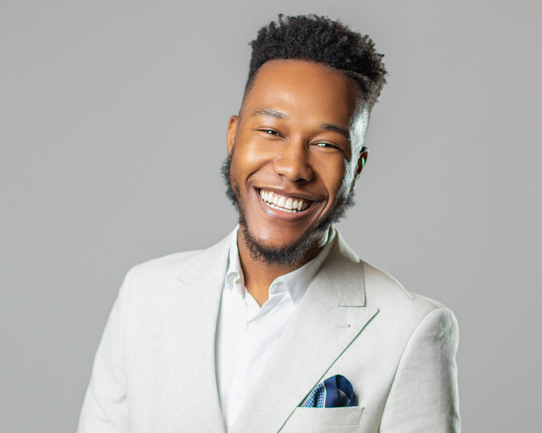 A smiling man with a beard and short curly hair, wearing a light-colored blazer over a white shirt, with a dark pocket square, against a plain light gray background.