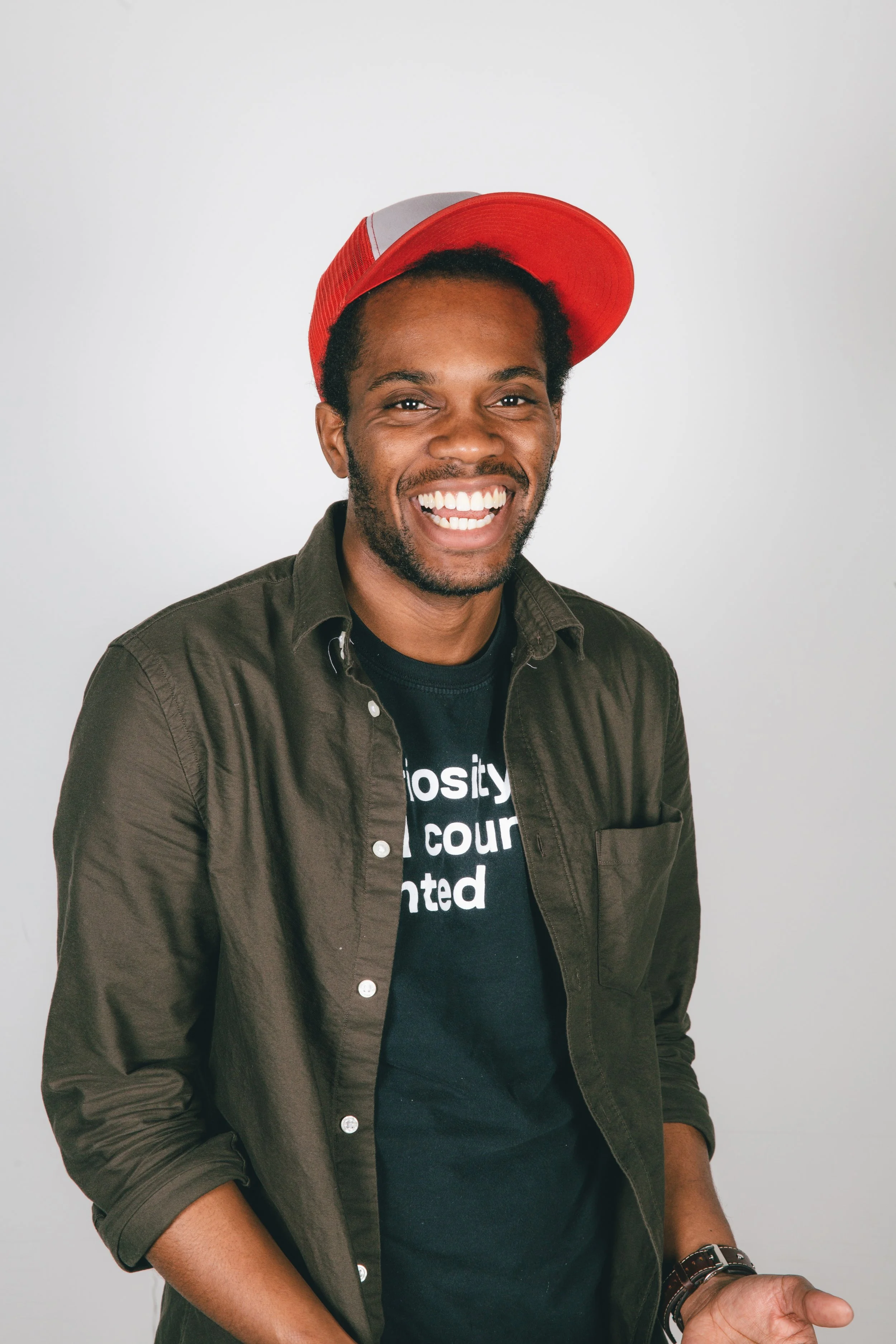 Smiling Black man wearing a red and gray baseball cap, green shirt, and a black t-shirt with white text, standing against a plain white background.