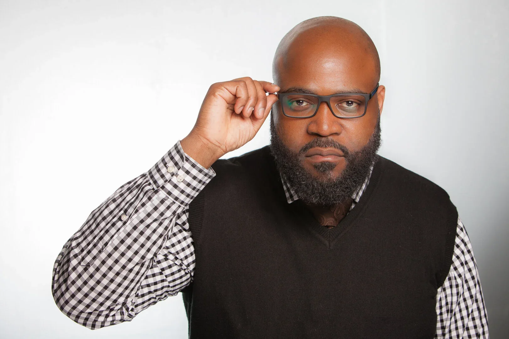 A man with a bald head and beard adjusting his glasses while wearing a checkered shirt and black vest, against a plain white background.