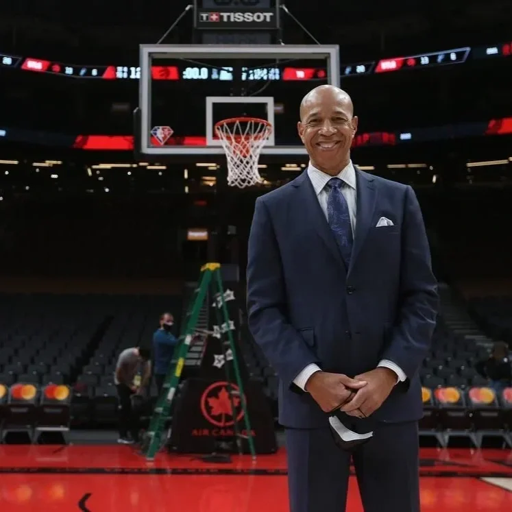 A man in a navy suit and tie standing on a basketball court, smiling, with a basketball hoop in the background.