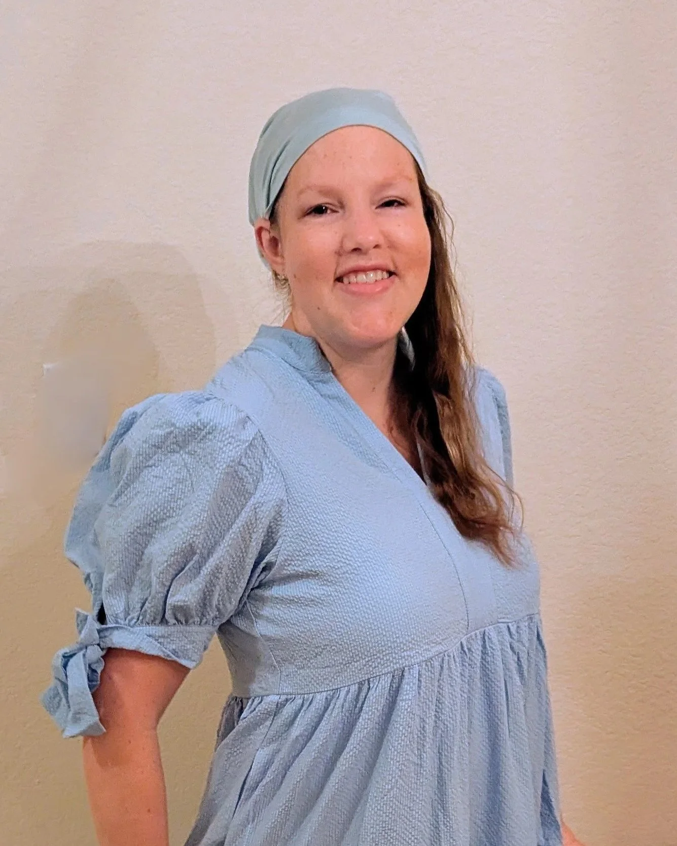 A woman with long brown hair wearing a light blue head covering and a blue dress suggests she might be a medical professional or nurse standing against a beige wall.