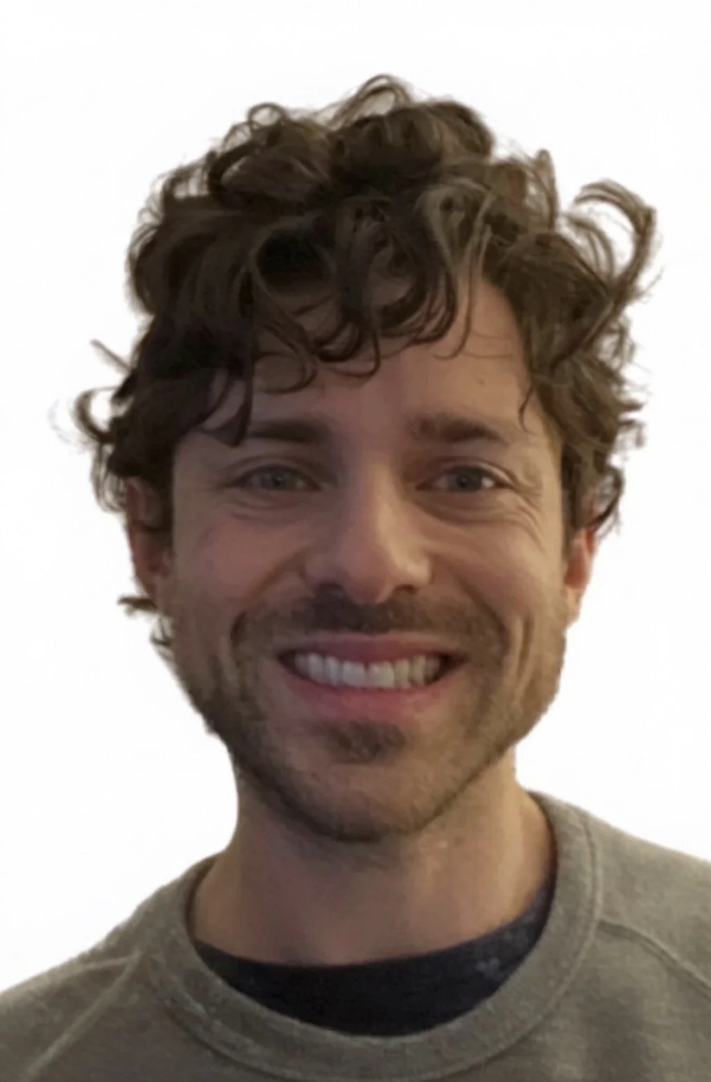 A smiling man with curly brown hair and light skin, wearing a gray shirt over a black undershirt, against a plain white background.