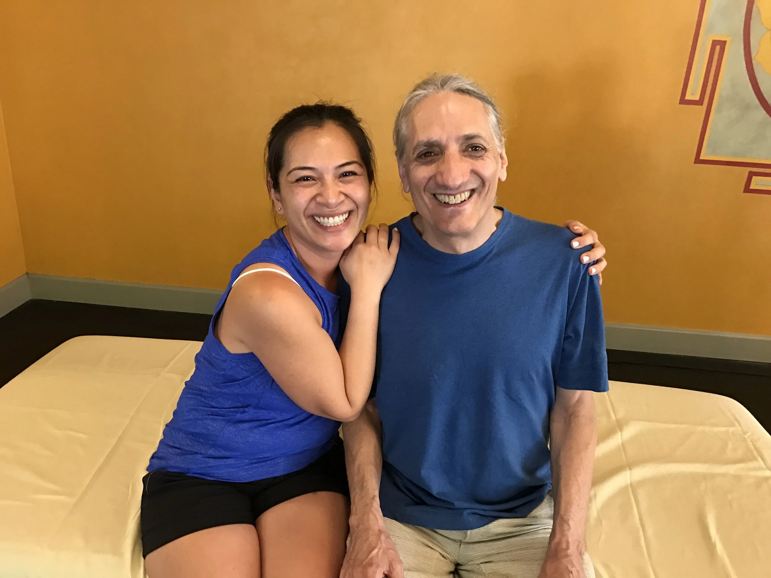 A woman with dark hair pulled back, wearing a blue sleeveless top and black shorts, smiling and hugging an older man with gray hair dressed in a blue shirt and beige pants, sitting on a yellow-covered bench against a yellow wall with a red and green decorative design.